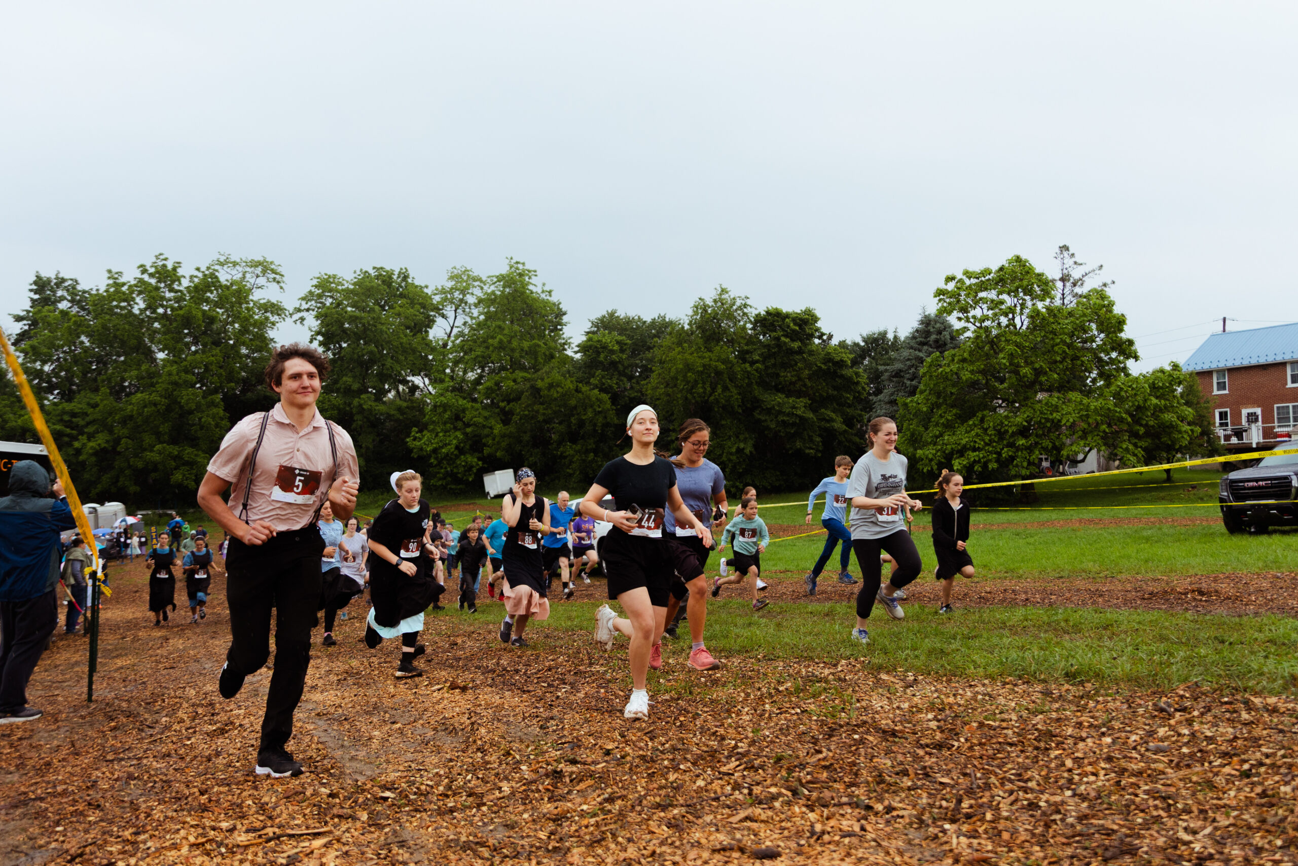 Participants in a community fun run race through a grassy field, showcasing a mix of ages and athletic wear, with trees and a barn in the background. The atmosphere is lively and energetic, capturing the spirit of local sports events.
