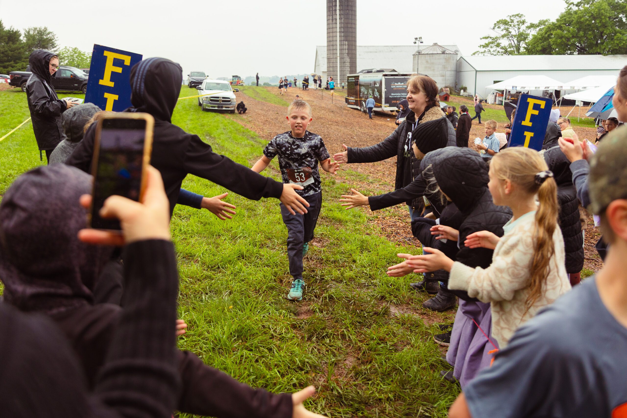 Child running through a cheering crowd at a youth race event, with supporters reaching out for high-fives and encouragement, set in a rural landscape with a cloudy sky.