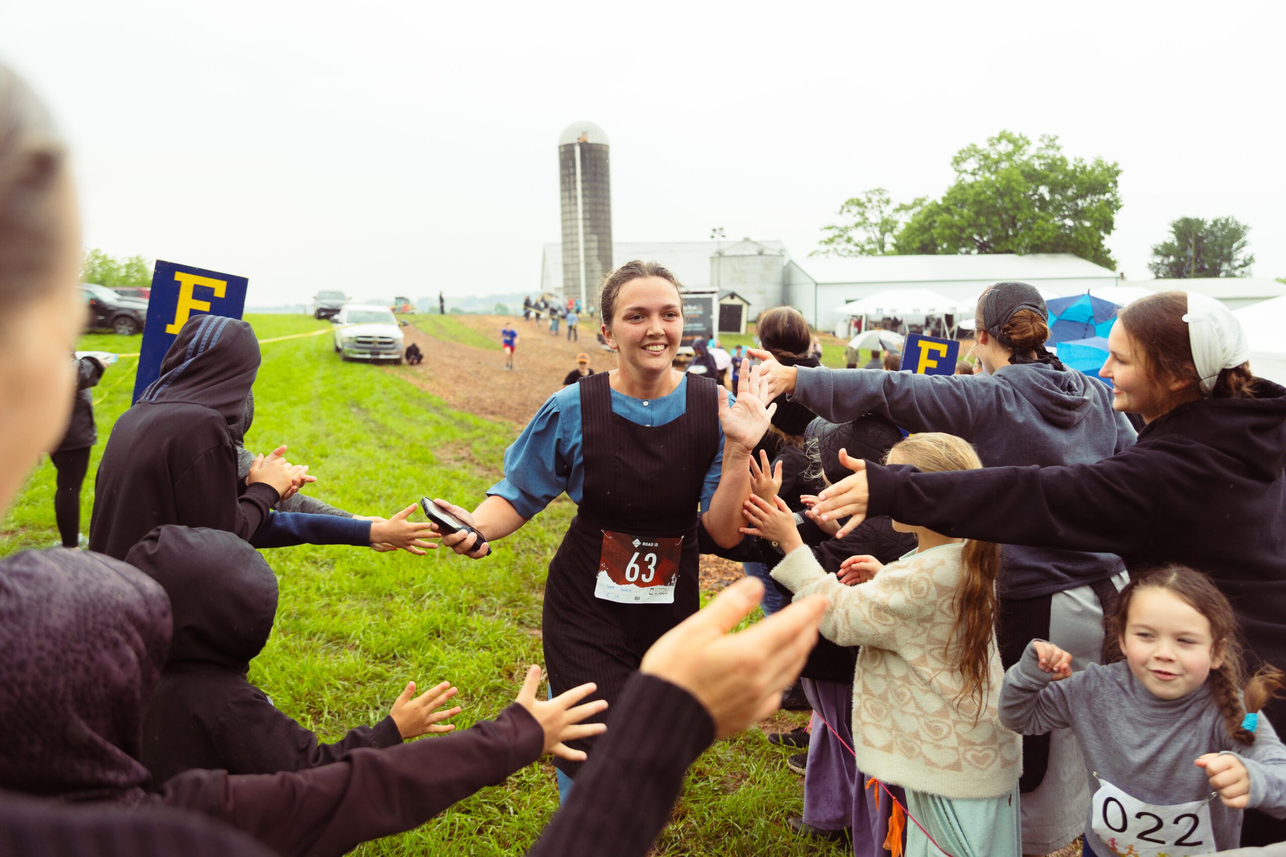 A smiling female runner in a black dress and blue shirt celebrates crossing the finish line at a rural event, surrounded by cheering supporters, including children and adults. A farm with silos and vehicles is visible in the background, indicating a community gathering focused on running or athletic competition.