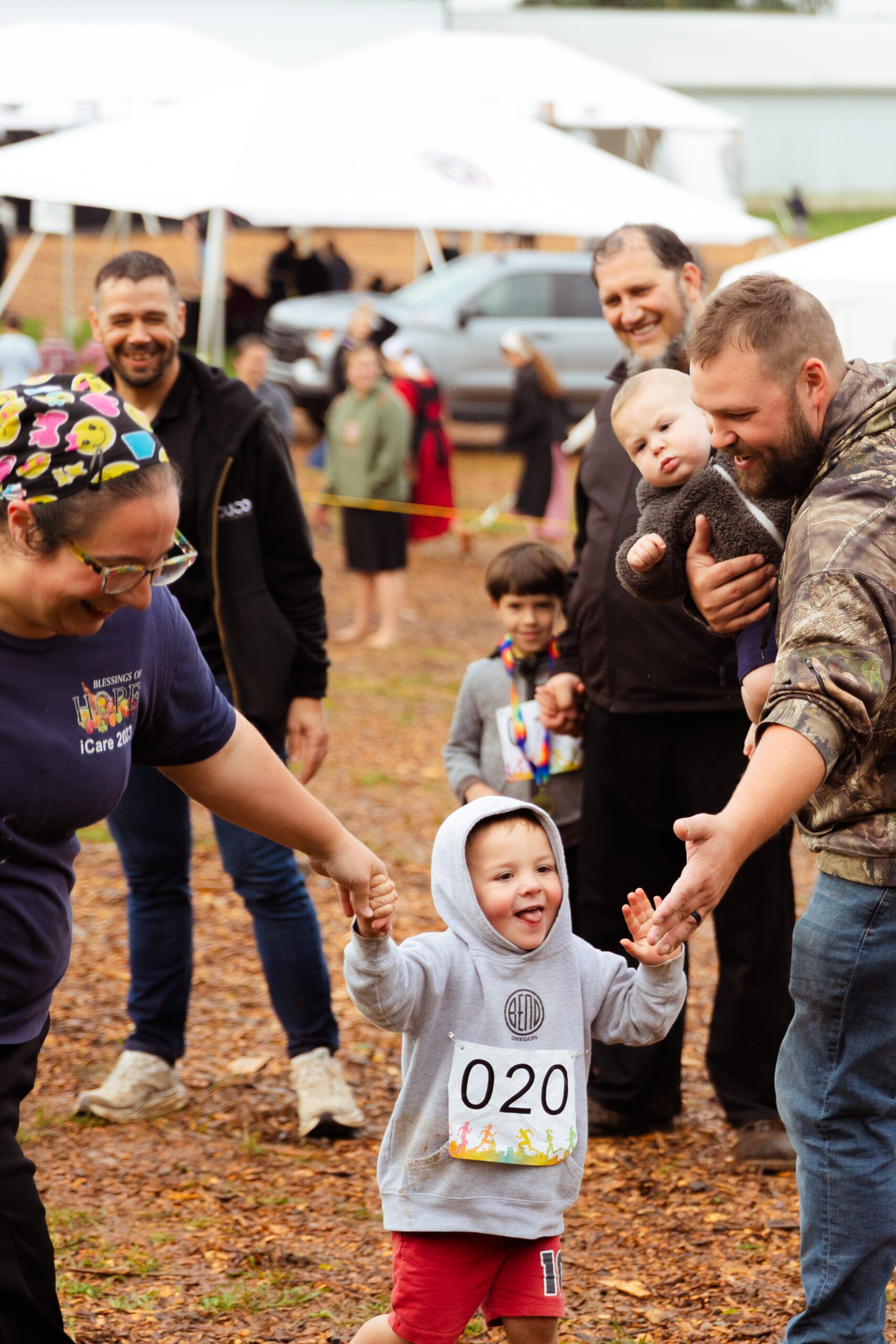 A joyful child in a gray hoodie and red shorts participates in a community event, holding hands with an adult while smiling. In the background, families gather under tents, creating a festive atmosphere.
