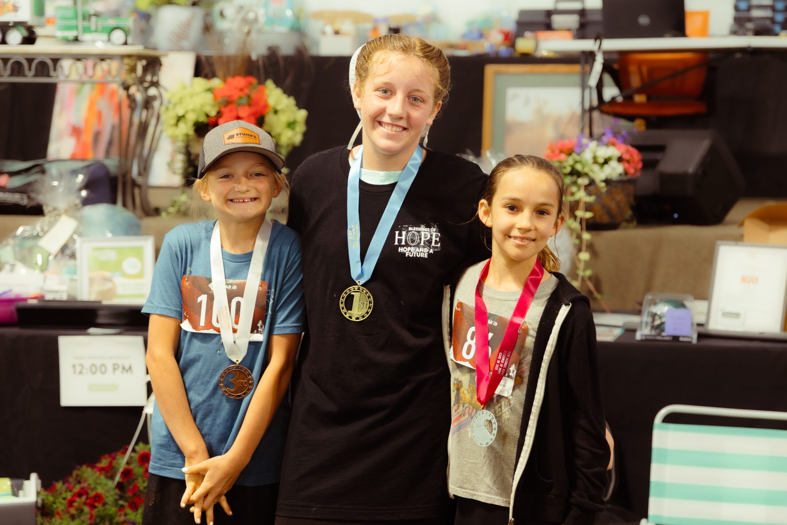 Three young athletes proudly display their medals at an event, with the girl in the center wearing a gold medal and the others showcasing bronze and silver. The background features colorful floral arrangements and event signage, highlighting their achievements in a friendly competition.