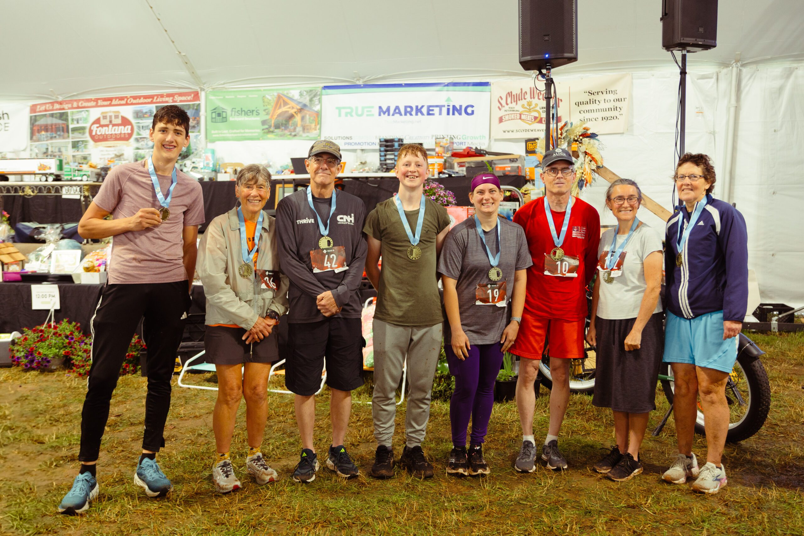 Group of eight medalists posing together after a running event, showcasing their medals in front of a tent filled with merchandise and floral arrangements.
