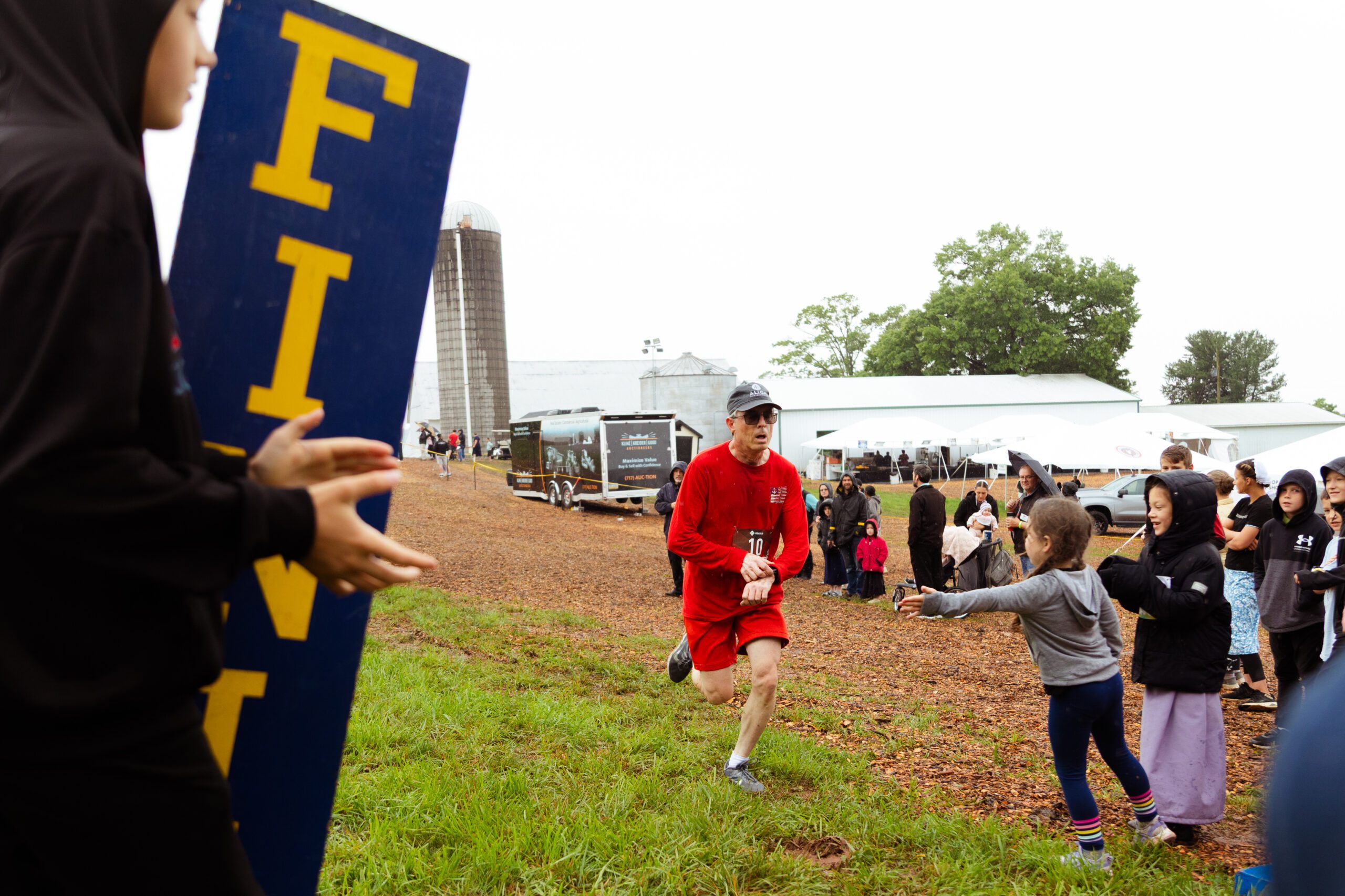 Runner in a red outfit crosses the finish line at a farm event, while spectators cheer and support from the sidelines. A gray sky looms overhead, indicating rainy weather, with a barn and tents visible in the background.