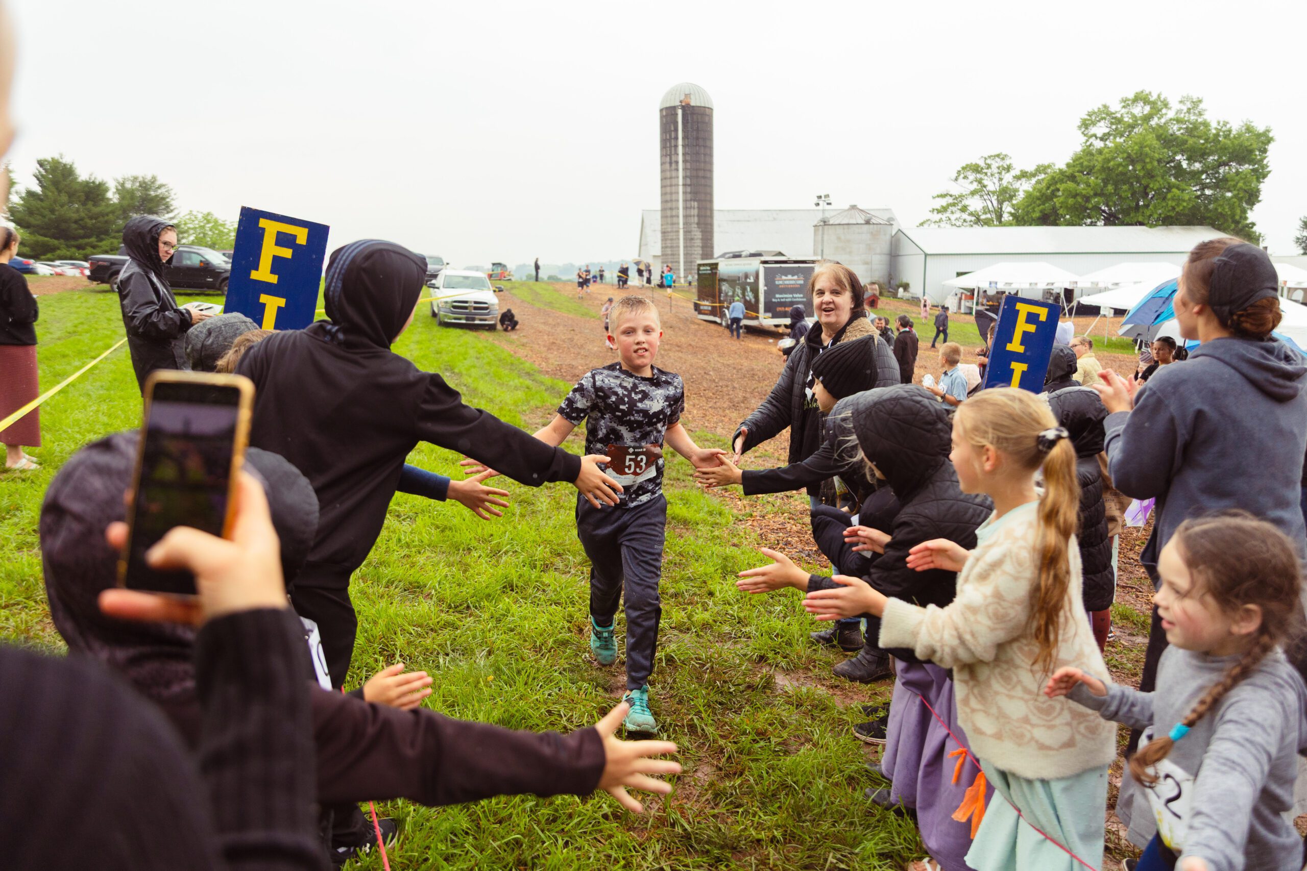 A young boy in a camouflage shirt runs through a cheering crowd at a rural running event, with supporters reaching out for high-fives. The background features a barn and vehicles, set in a grassy area under overcast skies.