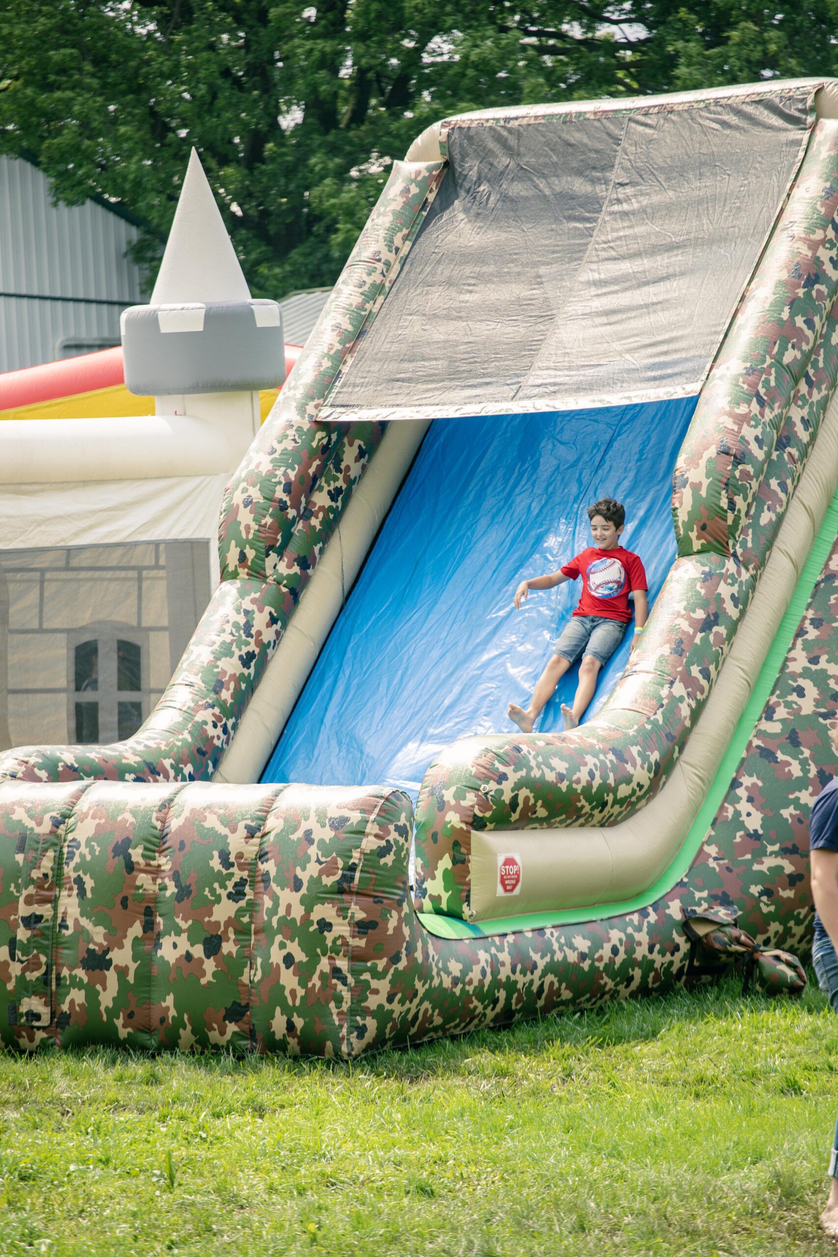 Child sliding down a large inflatable water slide with a camouflage design at an outdoor event, surrounded by green grass and other inflatable attractions in the background.
