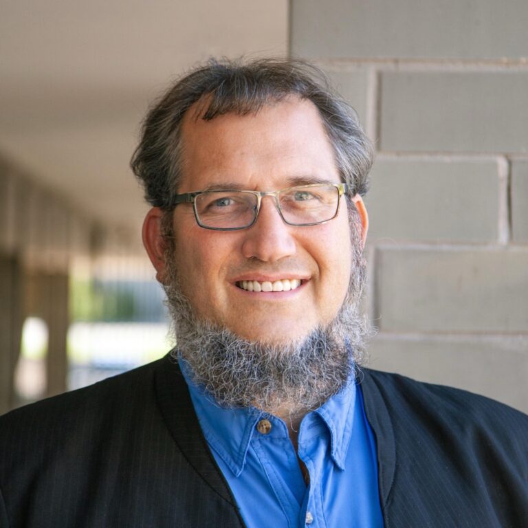 Smiling man with glasses and a beard, wearing a blue shirt and black jacket, standing in a corridor with gray brick walls. Suitable for profiles or professional introductions.