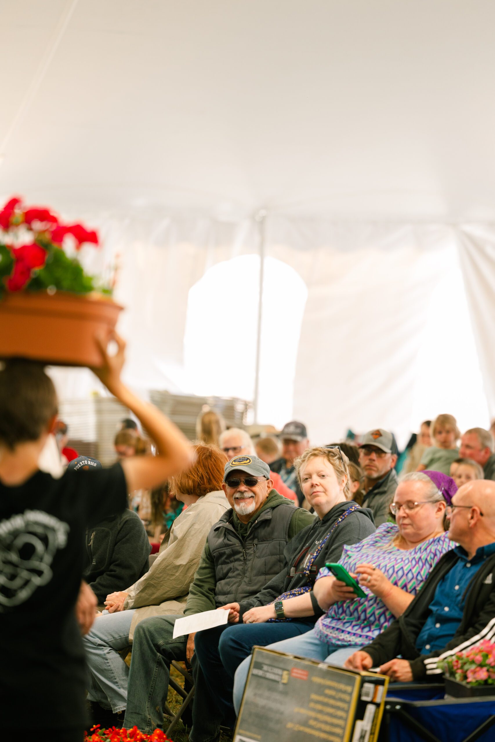 Audience at a gardening event seated under a tent, with a person holding a potted flower in the foreground. The attendees, including men and women of various ages, appear engaged and interested in the presentation.