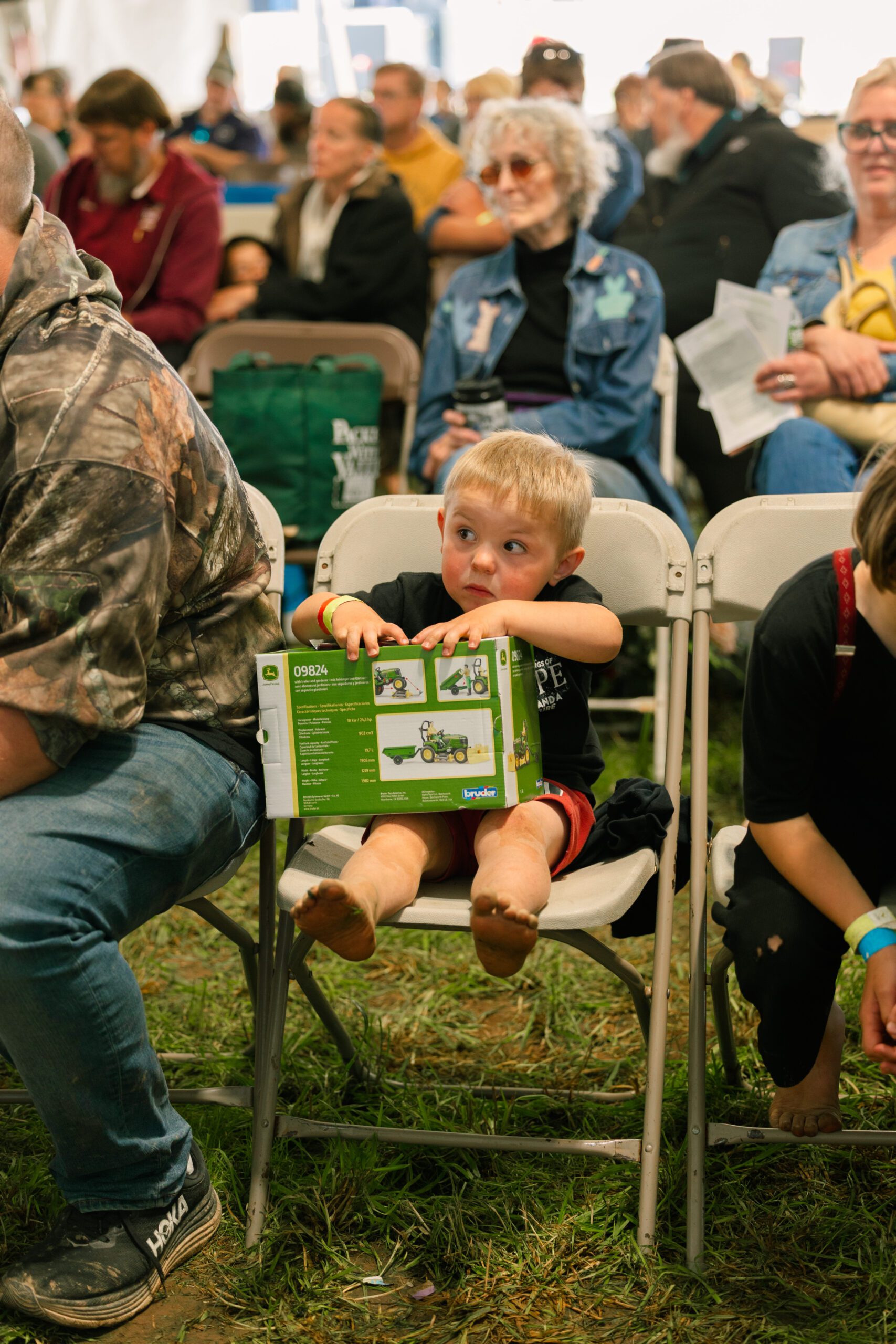 A young boy sitting on a folding chair at an outdoor event, holding a toy packaging while looking curiously at the crowd around him. The scene captures a casual family-friendly atmosphere with attendees engaged in various activities.