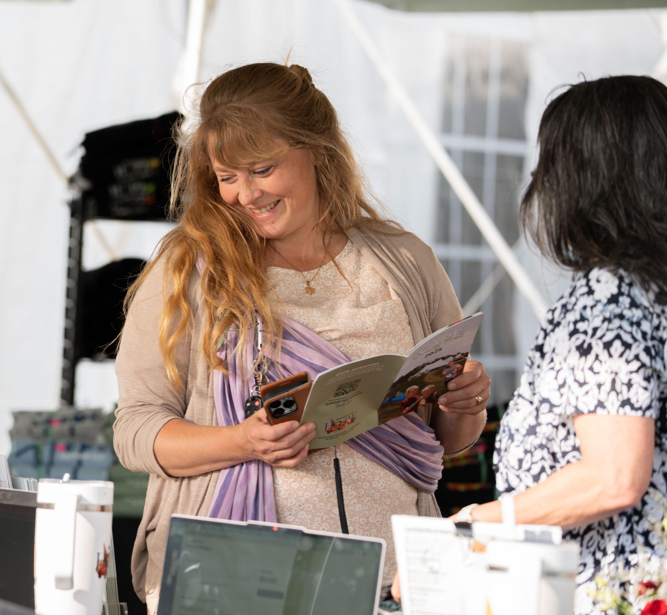 Woman smiling while reading a brochure at an outdoor event, with another person engaged in conversation nearby. Various items and displays are visible in the background, indicating a community gathering or market setting.