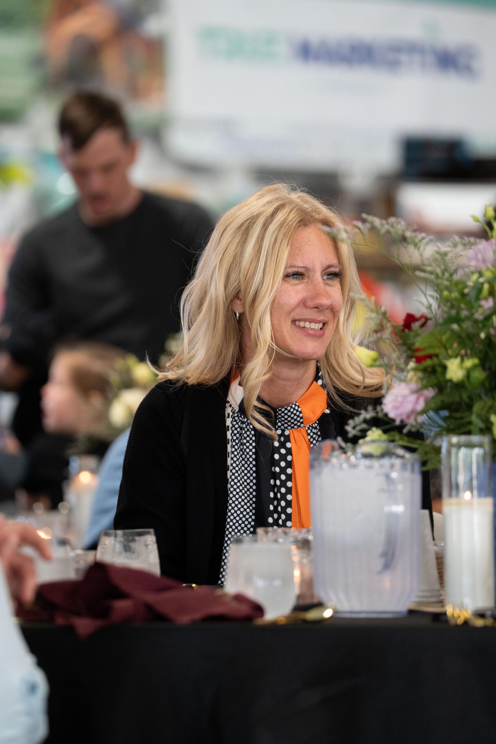 Smiling woman with long blonde hair wearing a polka dot blouse and orange scarf at a dining table adorned with flowers and candles, enjoying a social gathering in a festive setting.