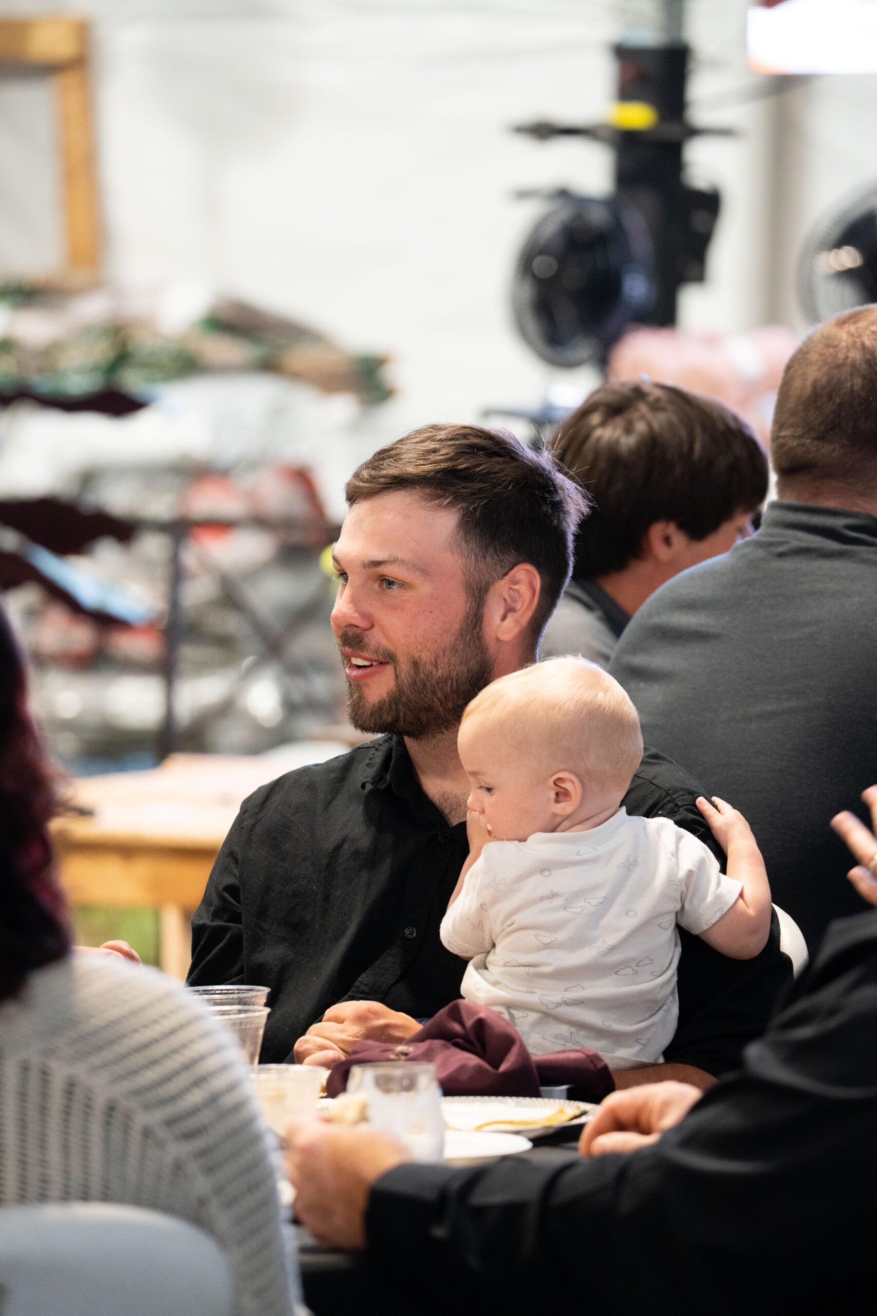 A young man with a beard sits at a table holding a baby in a cozy indoor setting, surrounded by other diners and soft lighting.