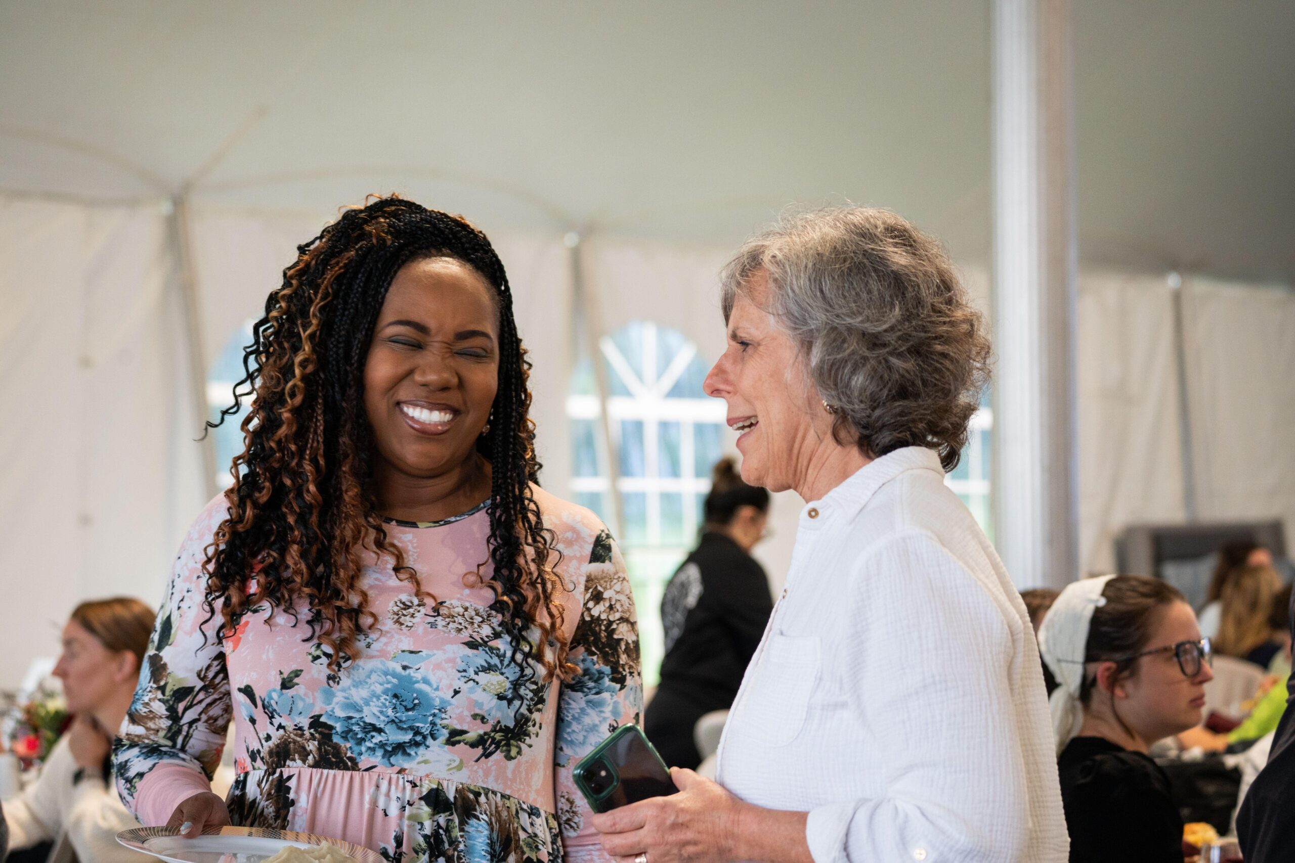 Two women smiling and chatting at a social gathering under a tent, with one woman holding a plate of food and the other holding a phone. The scene captures a warm, friendly atmosphere.