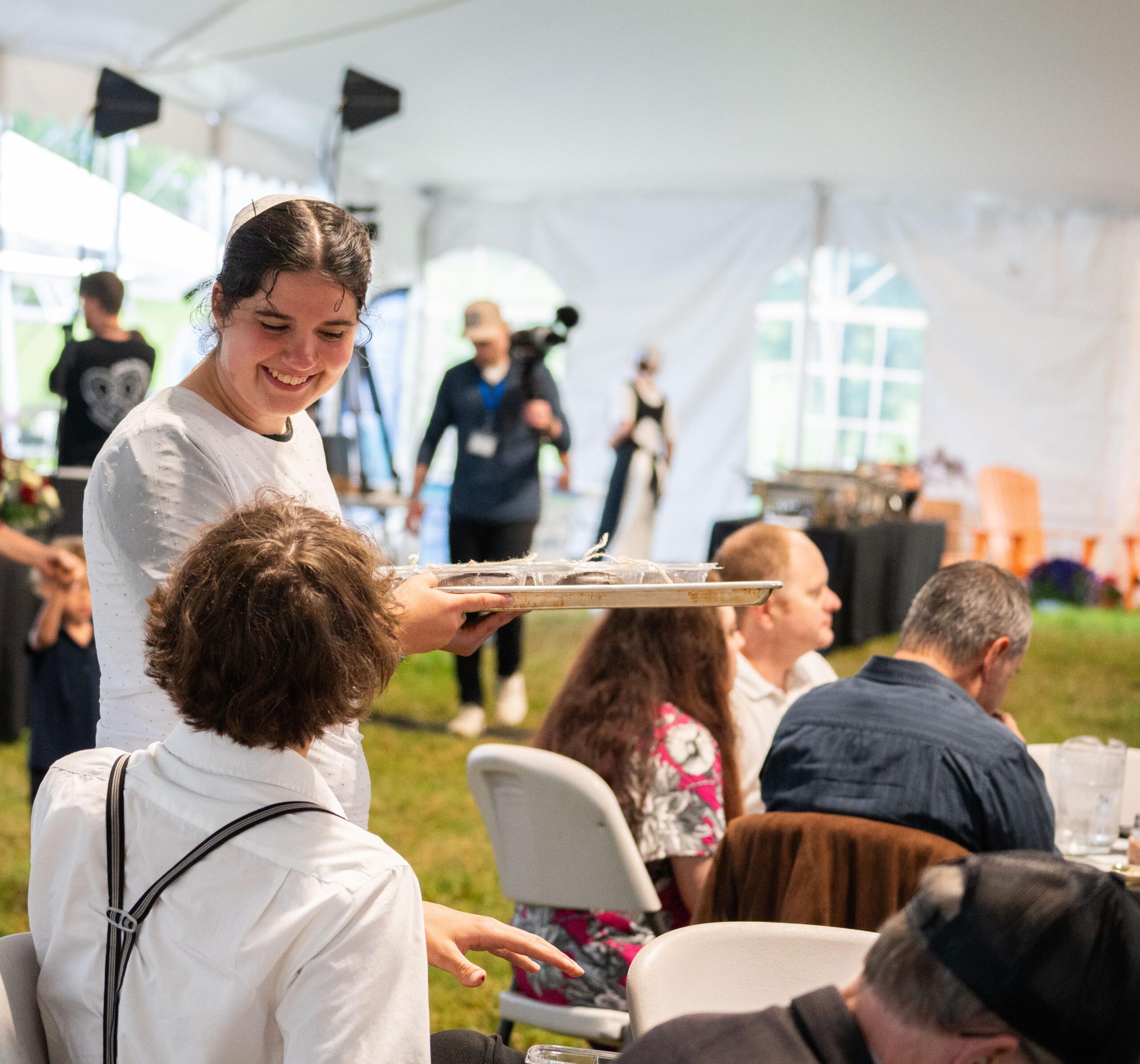 A waitress serves food on a tray to a guest at a tented outdoor event, with other attendees enjoying their meals in the background.