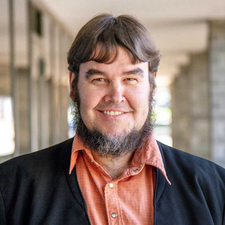 Smiling man with a beard wearing an orange shirt and black jacket, standing in a well-lit corridor.