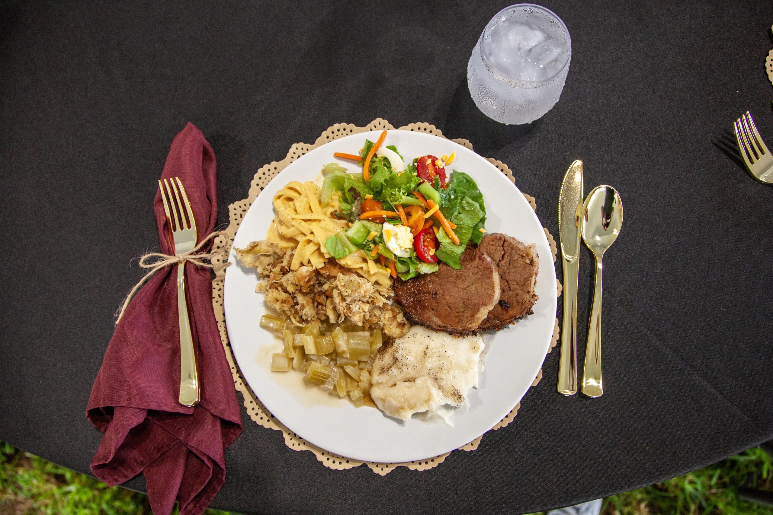 A beautifully arranged plate featuring a variety of dishes including sliced beef, creamy mashed potatoes, seasoned vegetables, pasta, and a fresh salad with mixed greens and cherry tomatoes, set on an elegant table with gold utensils and a glass of water.