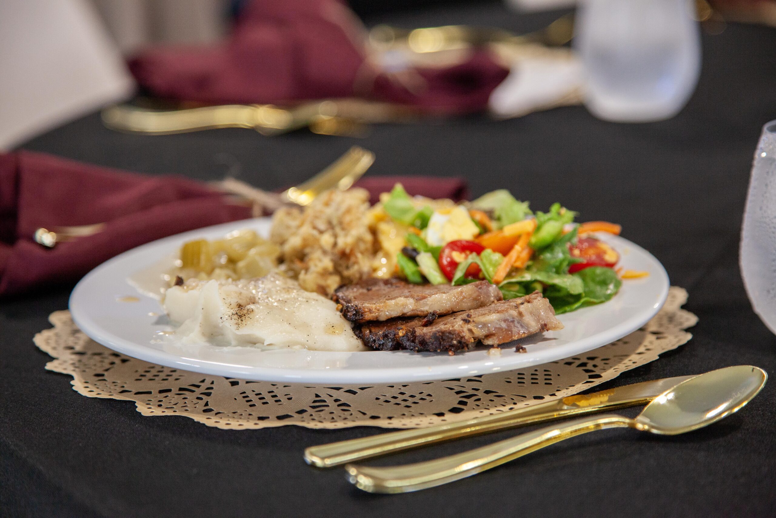 A beautifully arranged plate featuring sliced meat, creamy mashed potatoes, stuffing, green beans, and a fresh salad with cherry tomatoes and mixed greens, set on an elegant table with gold utensils and a lace doily.