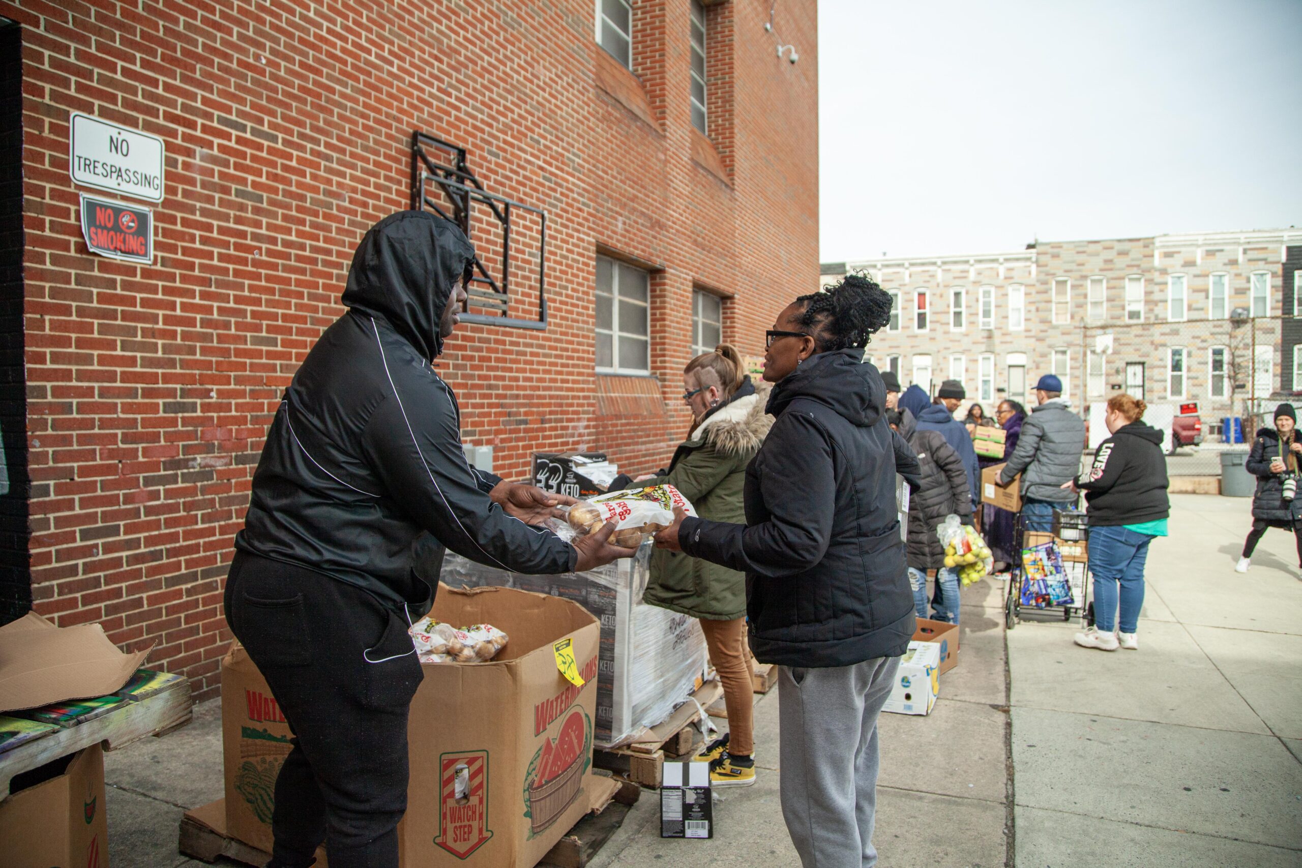 Community members participating in a food distribution event, with volunteers handing out groceries outside a brick building. Boxes of food are visible, and people are lined up receiving assistance in a supportive environment.
