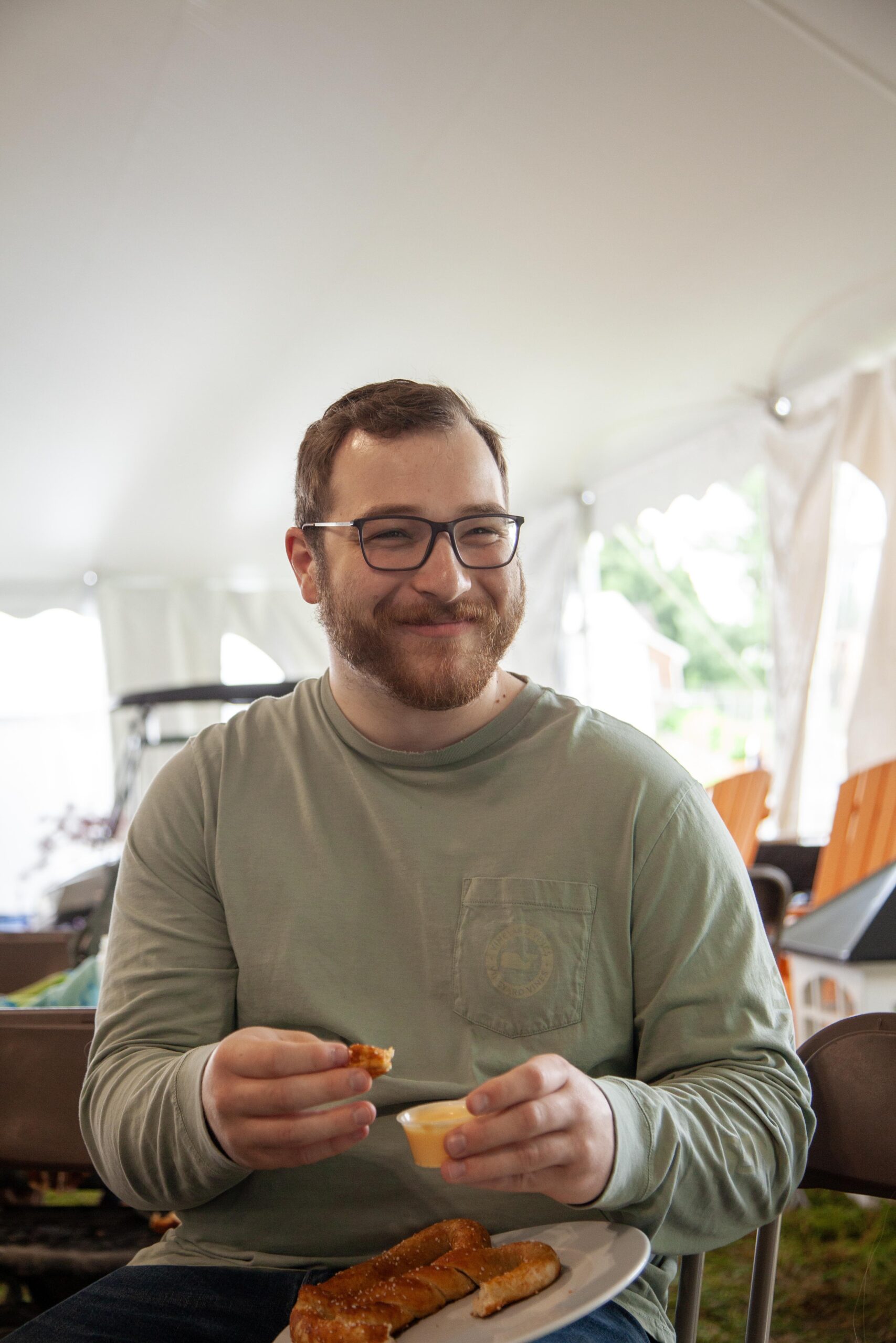 Smiling man with glasses enjoying a plate of food, including a pretzel and dipping sauce, seated under a tent at an outdoor event.