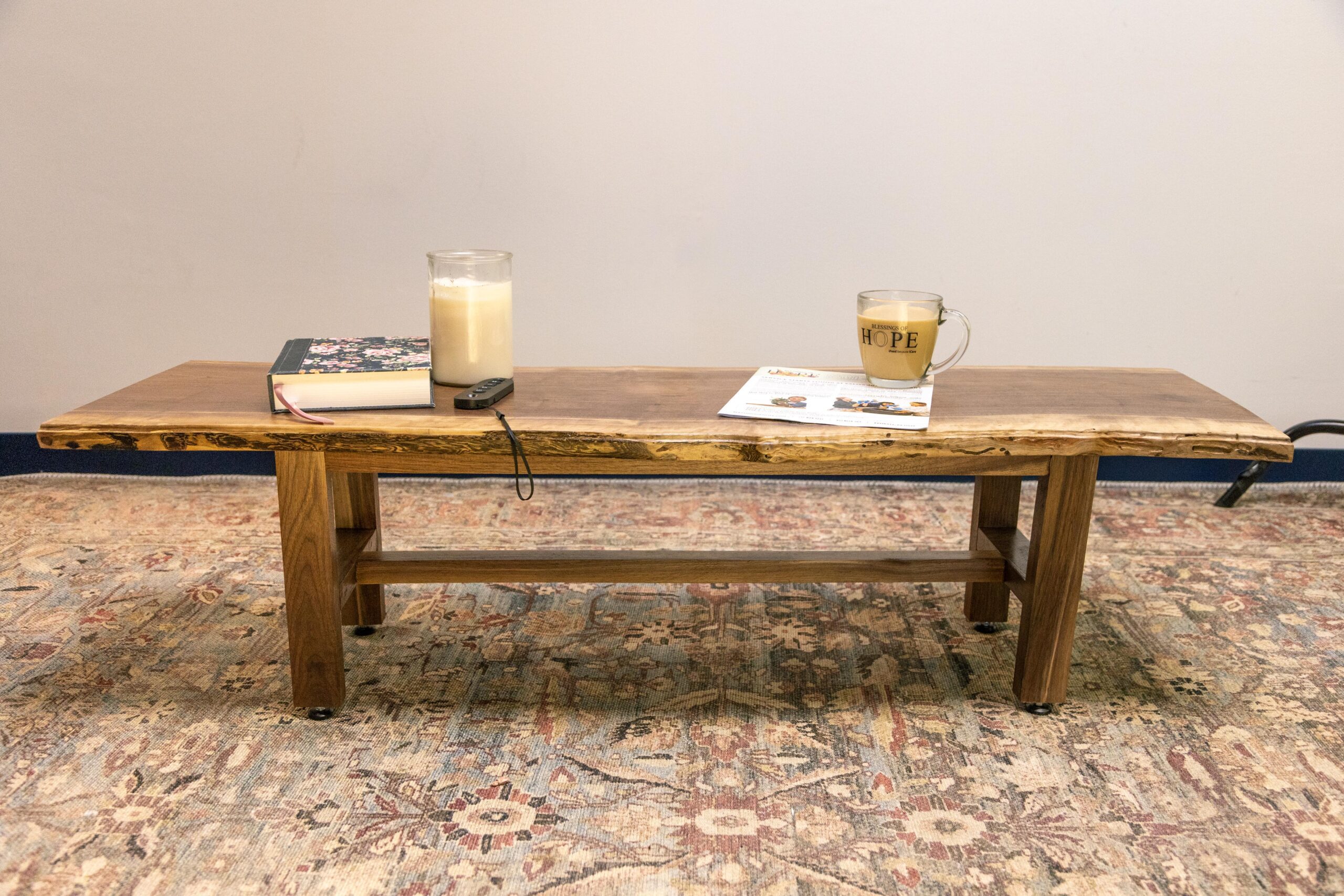 Rustic wooden coffee table with a live edge, featuring a candle, a notebook, a remote control, and a mug on top, set on a patterned area rug.