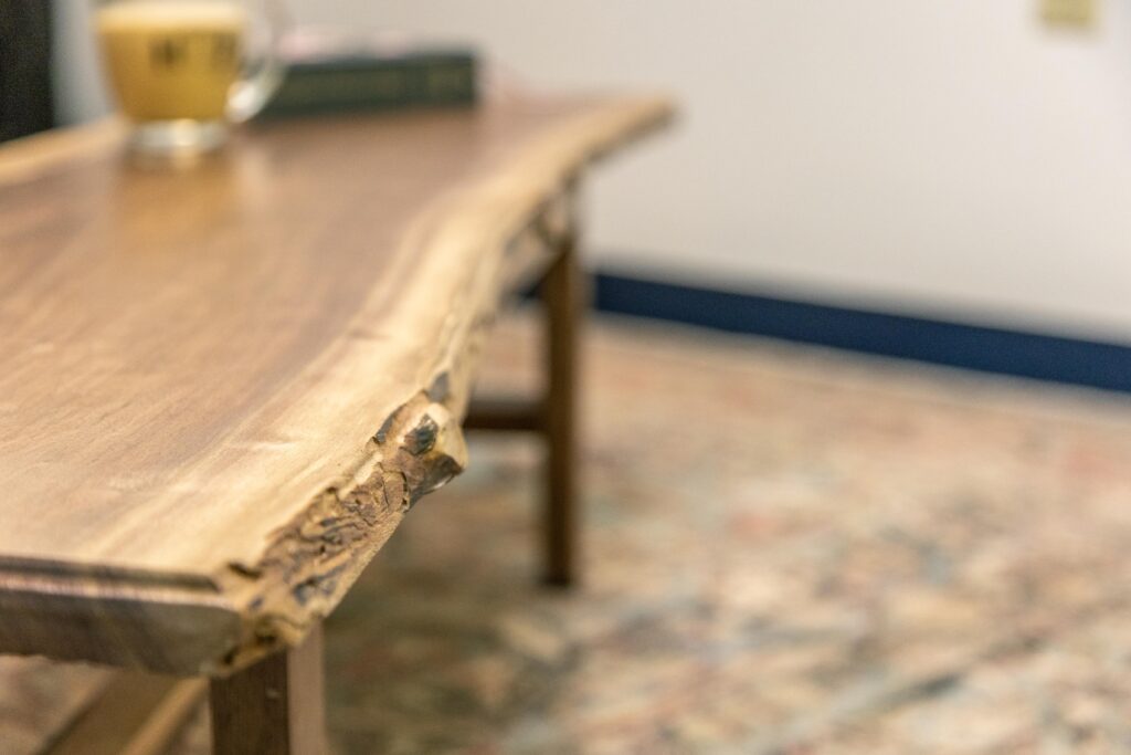 Close-up of a natural wood coffee table featuring a live edge, highlighting its unique grain and texture, with a blurred background showcasing a patterned rug and a glass of beverage.
