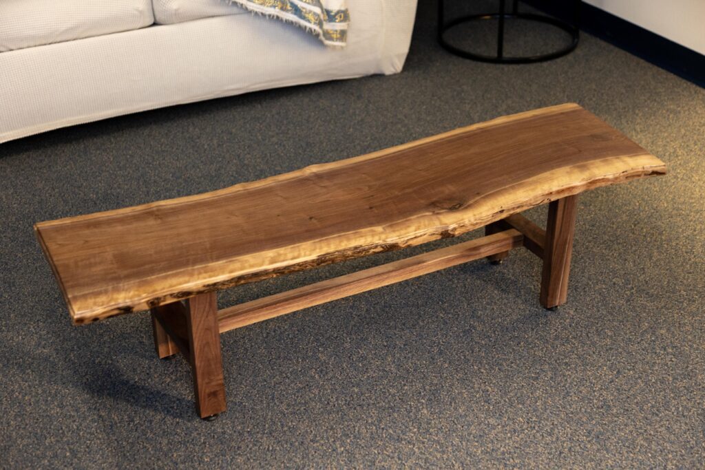 Natural wood bench with a live edge design, featuring sturdy wooden legs, set in a cozy living room environment with a sofa in the background. Ideal for rustic or contemporary home decor.