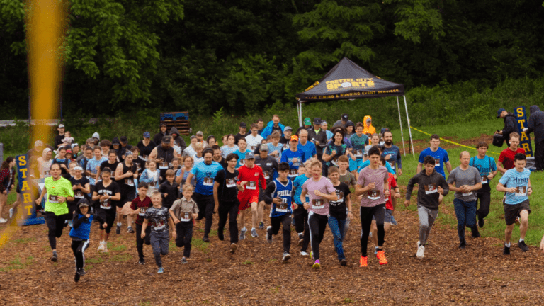 Participants sprinting at the start of a community fun run event, showcasing a diverse group of runners on a wooded trail surrounded by greenery and a start line banner.