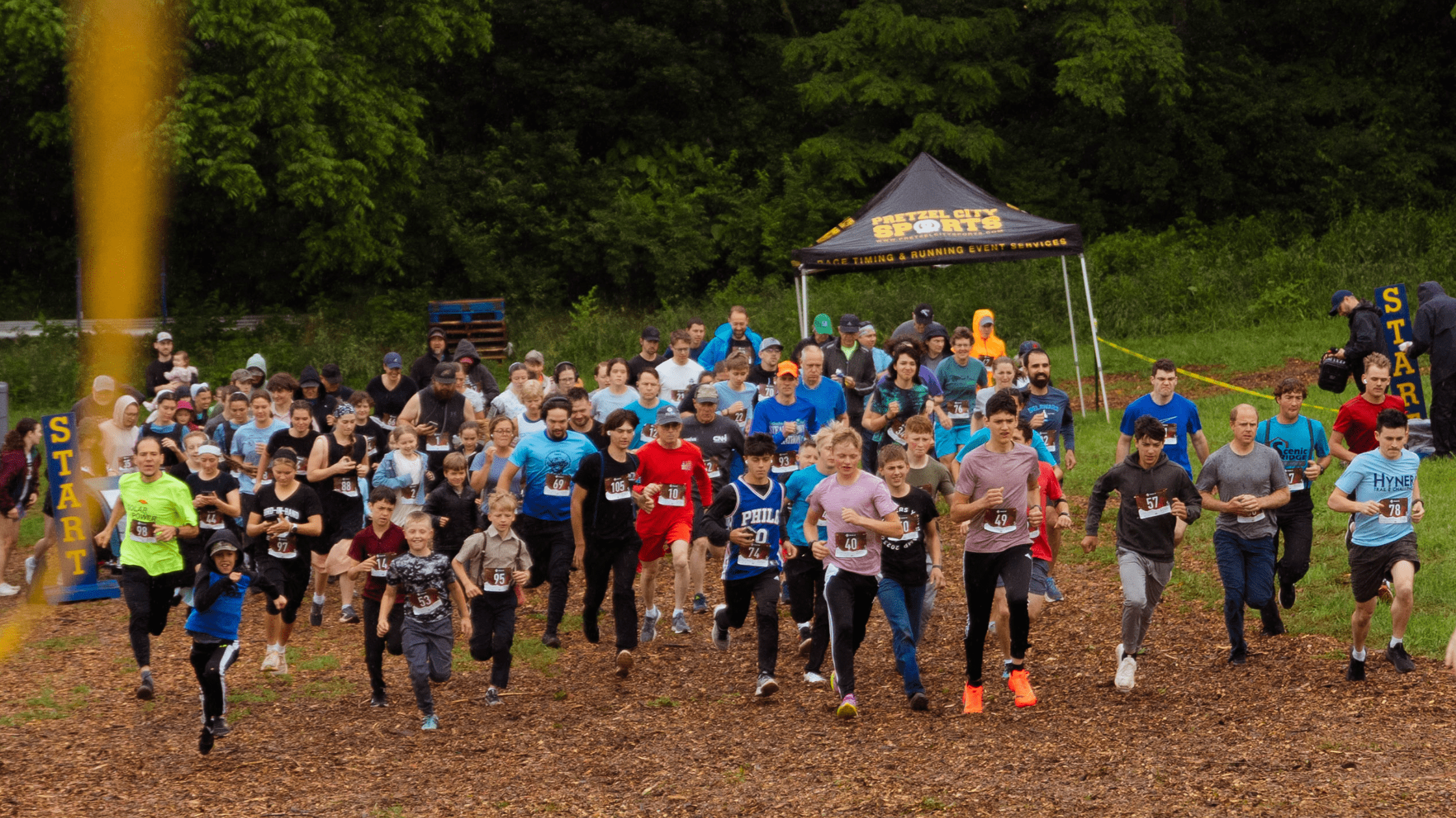 Participants sprinting at the start of a community fun run event, showcasing a diverse group of runners on a wooded trail surrounded by greenery and a start line banner.