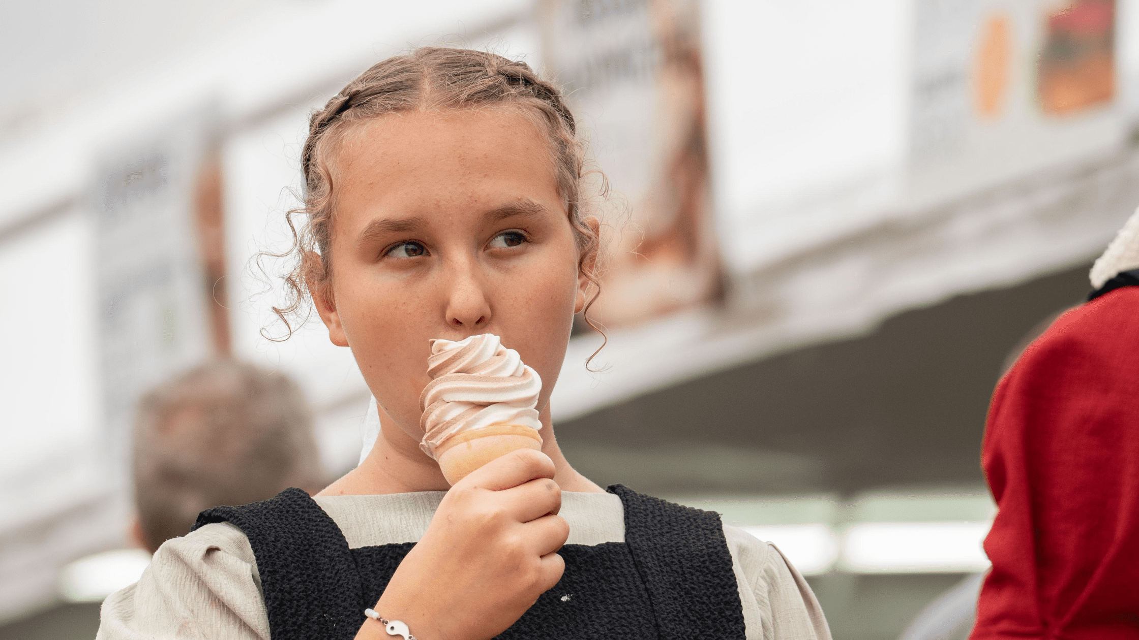 Young girl enjoying a soft-serve ice cream cone with a chocolate and vanilla swirl, wearing a traditional outfit, at a fair or outdoor event.