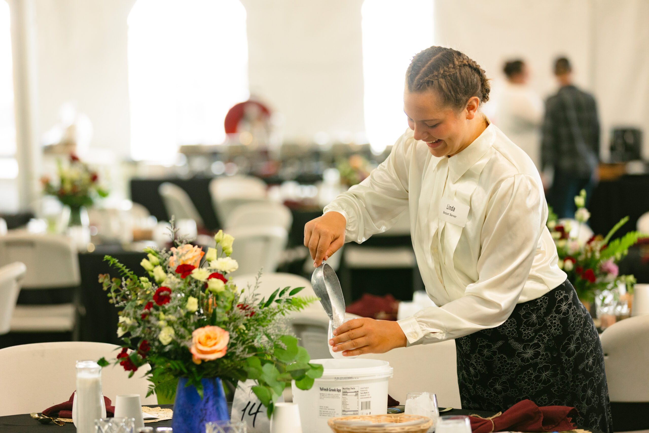 A server in a white blouse and black skirt prepares a dining table with floral centerpieces at an event venue, showcasing elegant table settings and a welcoming atmosphere.
