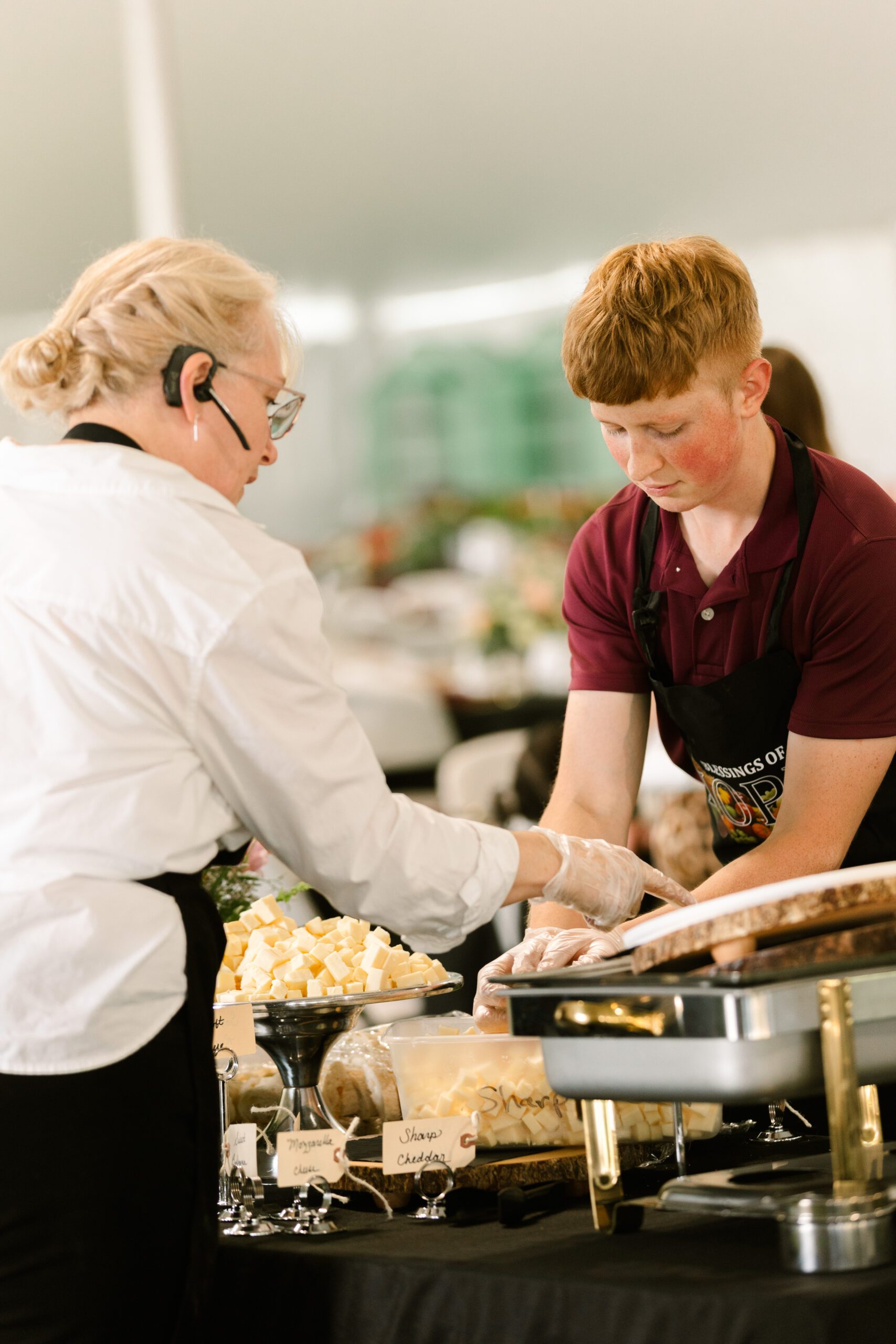 Catering staff serve cheese at an outdoor event, with a woman in a white shirt and headset assisting a young man in a maroon shirt. The table features a variety of cheeses labeled for guests.