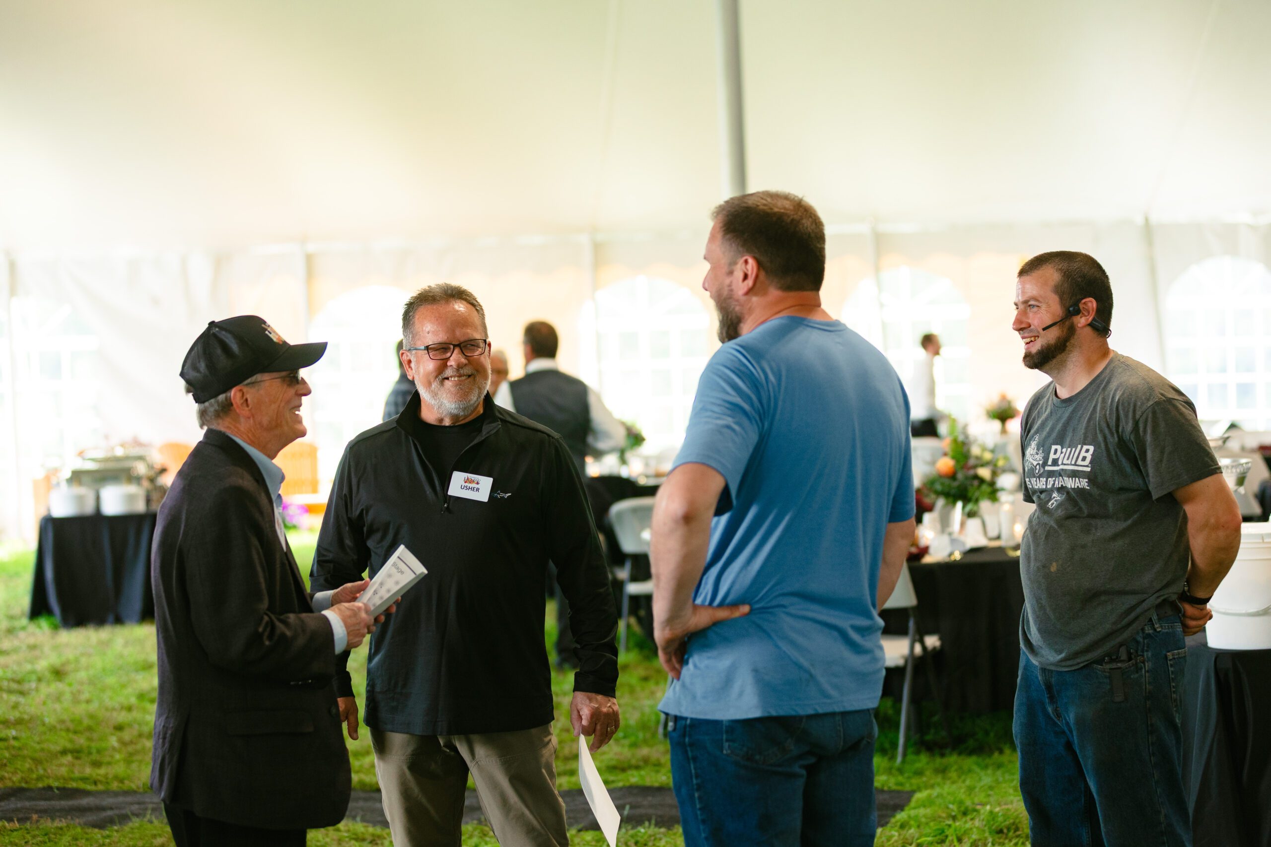 Group of four men engaging in conversation at an outdoor event under a tent, with tables set for a gathering. The setting features floral arrangements and a buffet in the background.