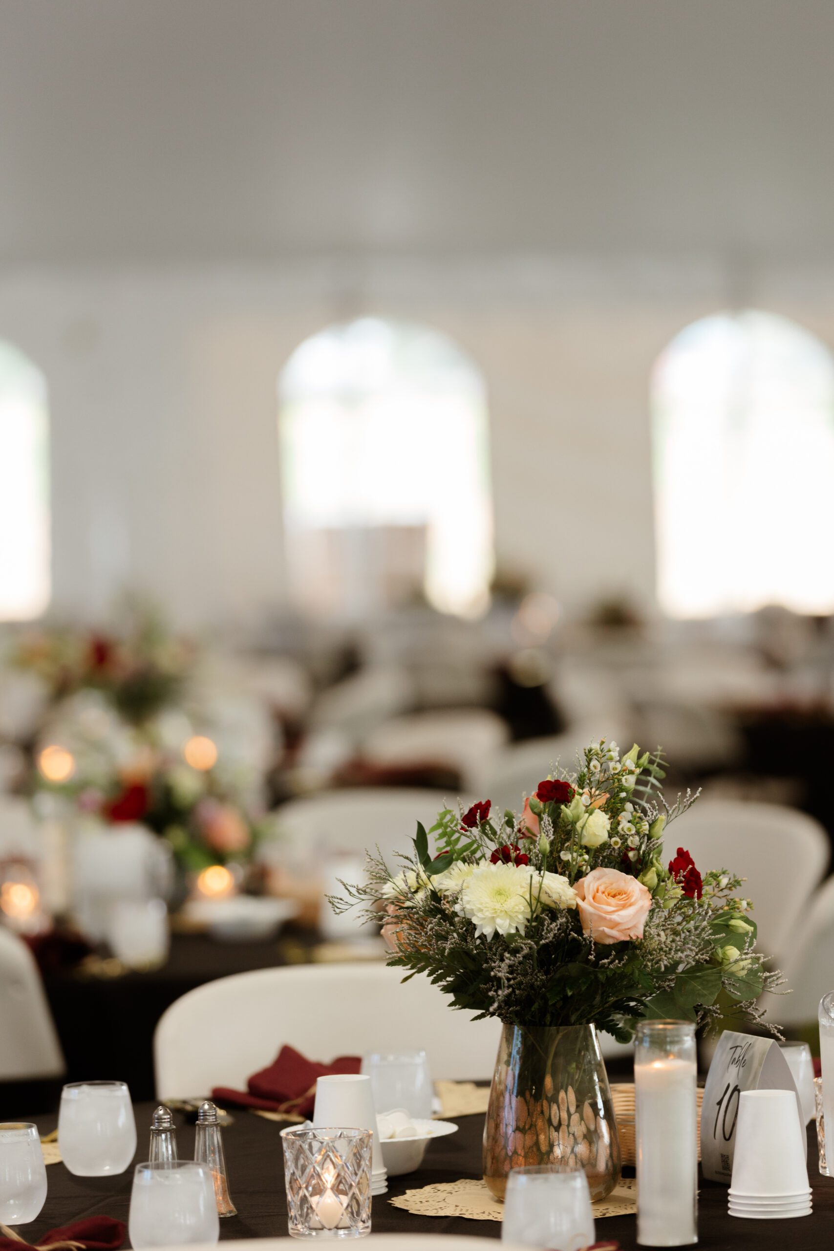 Elegant table setting at a tented event featuring a floral centerpiece with roses and greenery, surrounded by white chairs and soft candlelight.