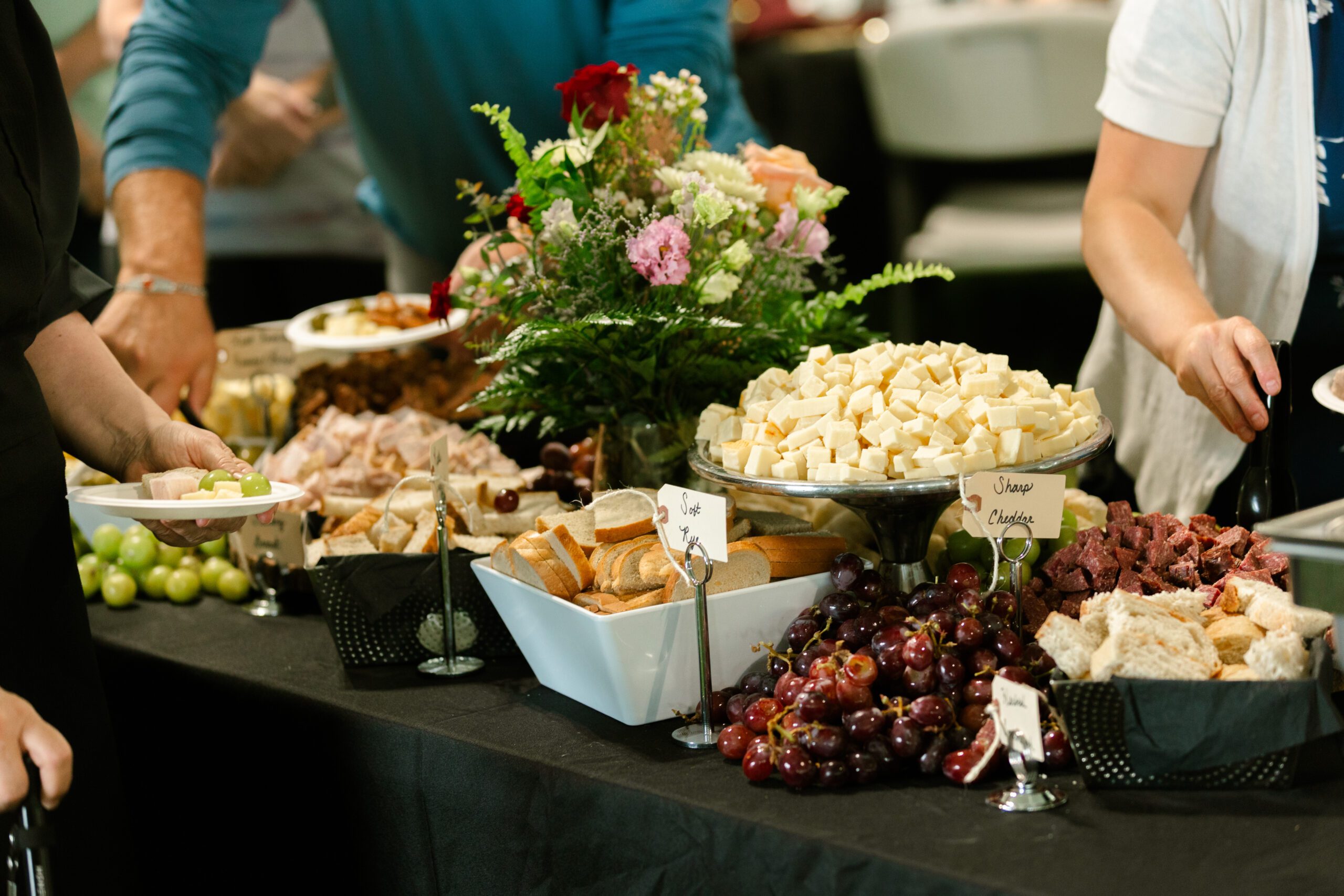A buffet table featuring a variety of food options, including slices of ham, cubes of sharp cheddar cheese, assorted breads, and fresh grapes. Floral arrangements add a decorative touch, while guests serve themselves from plates.