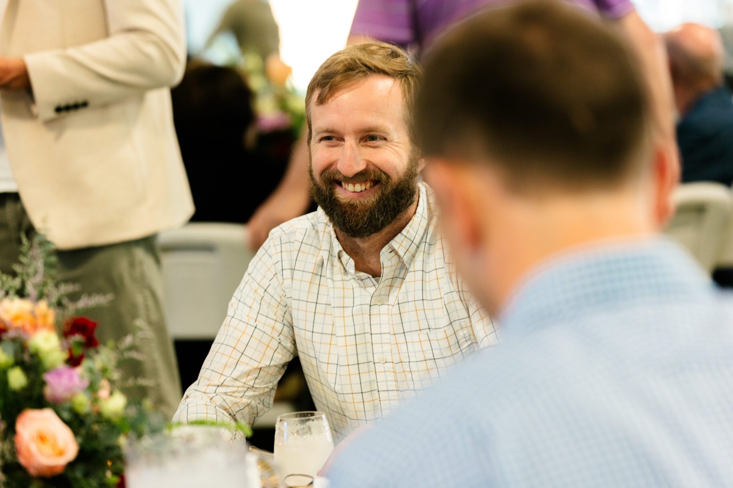 Smiling man with a beard sitting at a table during a social gathering, surrounded by floral arrangements and other attendees.