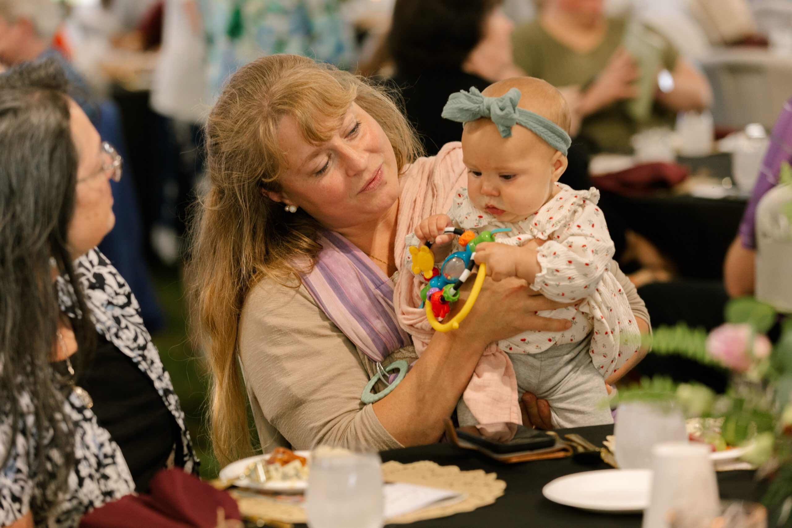 Woman holding a baby with a colorful toy at a social gathering, surrounded by people at tables, showcasing a warm family moment.