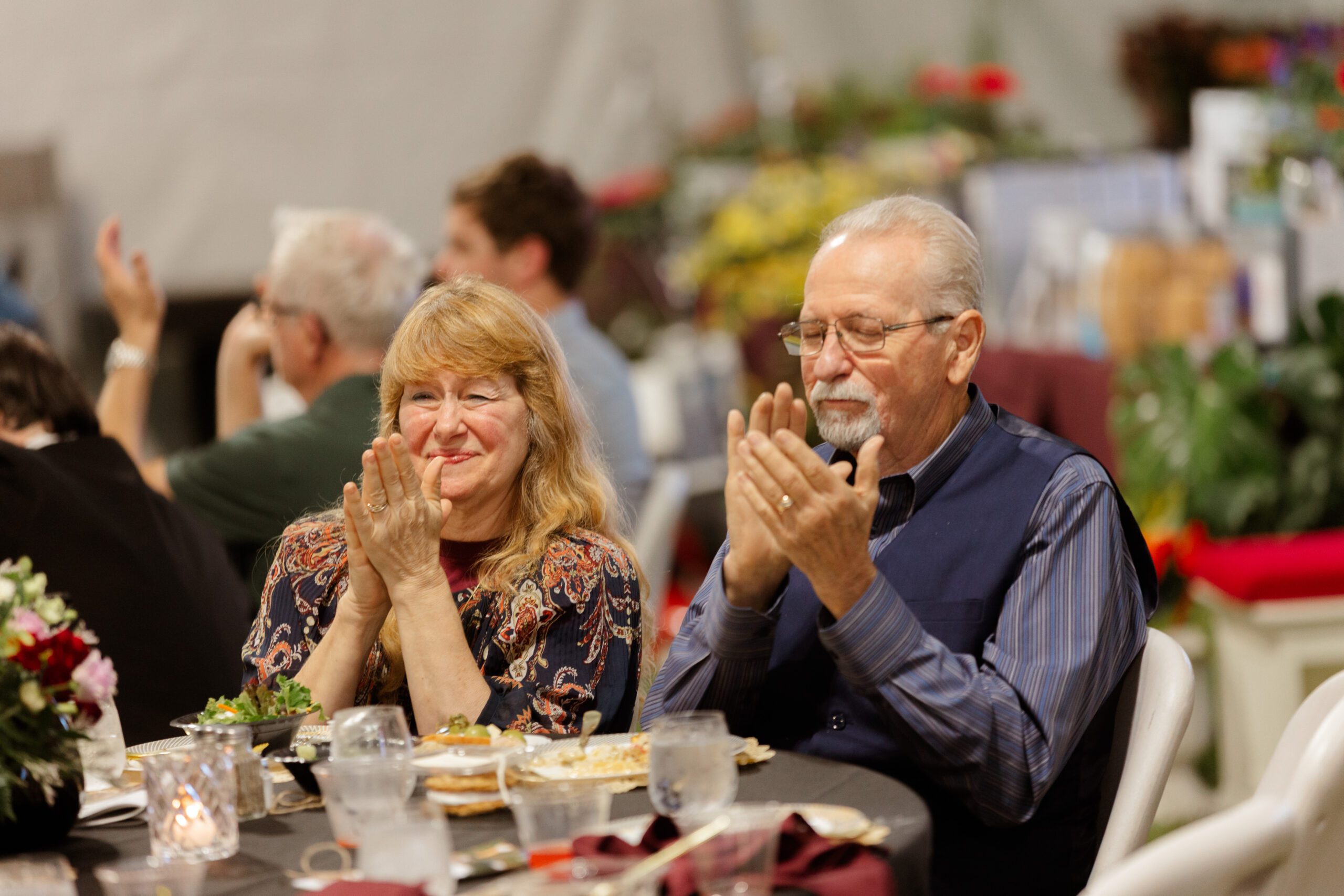 Couple enjoying a celebratory event, clapping and smiling at the table, surrounded by friends and festive decorations.