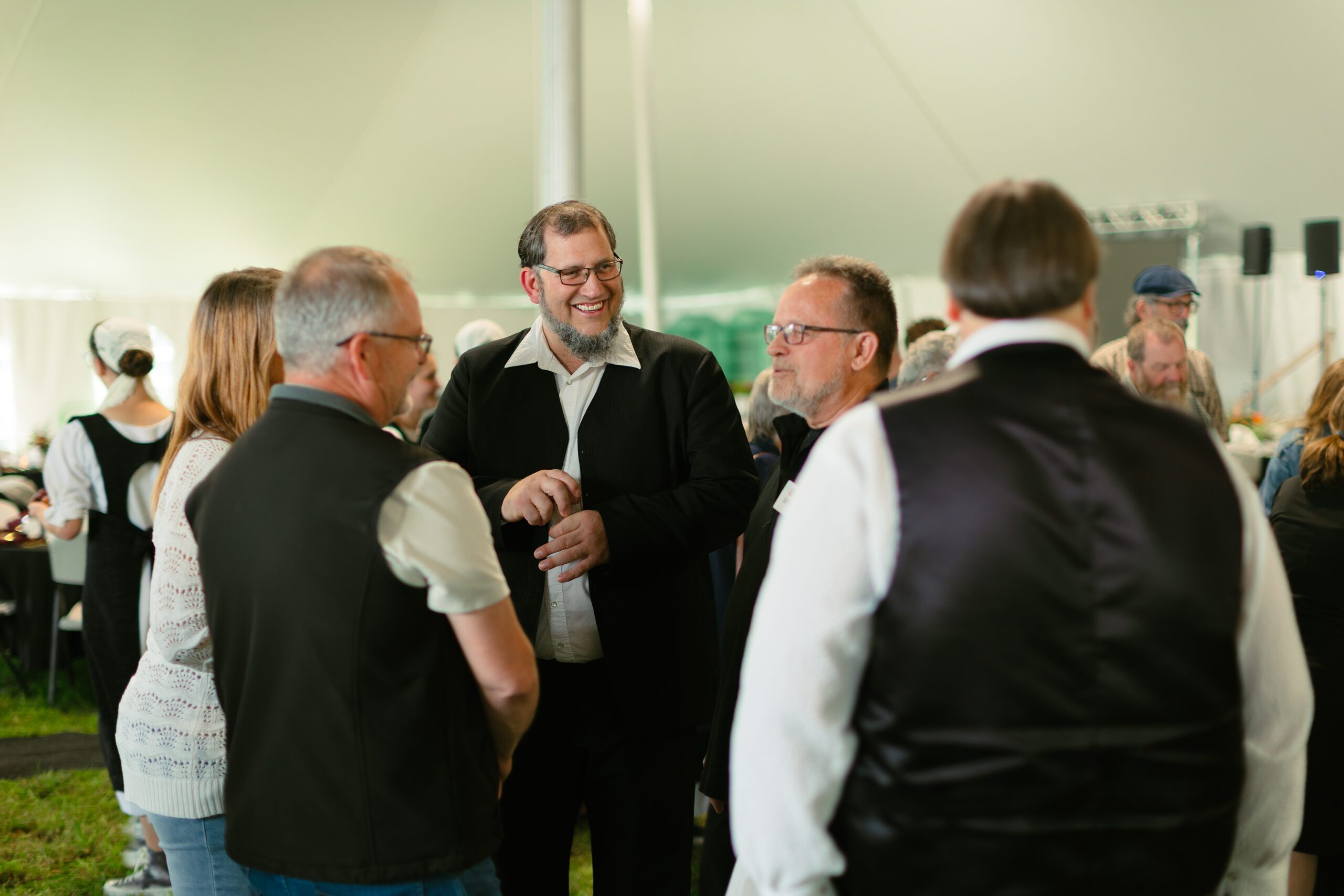 Group of people engaging in conversation at a social event under a tent, showcasing a friendly atmosphere with individuals dressed in semi-formal attire.
