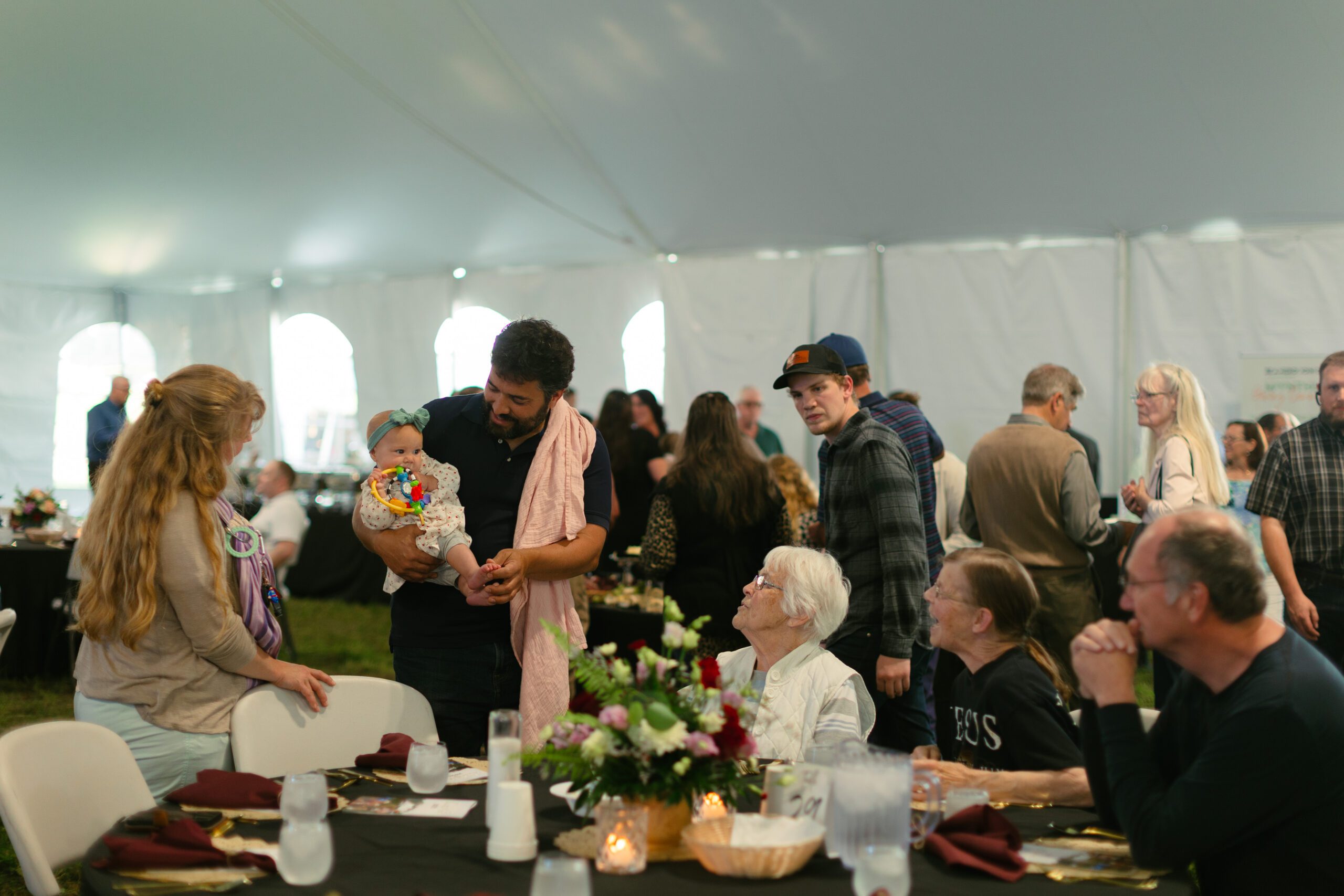 Family gathering under a tent with a diverse group of people, including adults and a baby, enjoying a meal and conversation, surrounded by festive table settings and floral arrangements.