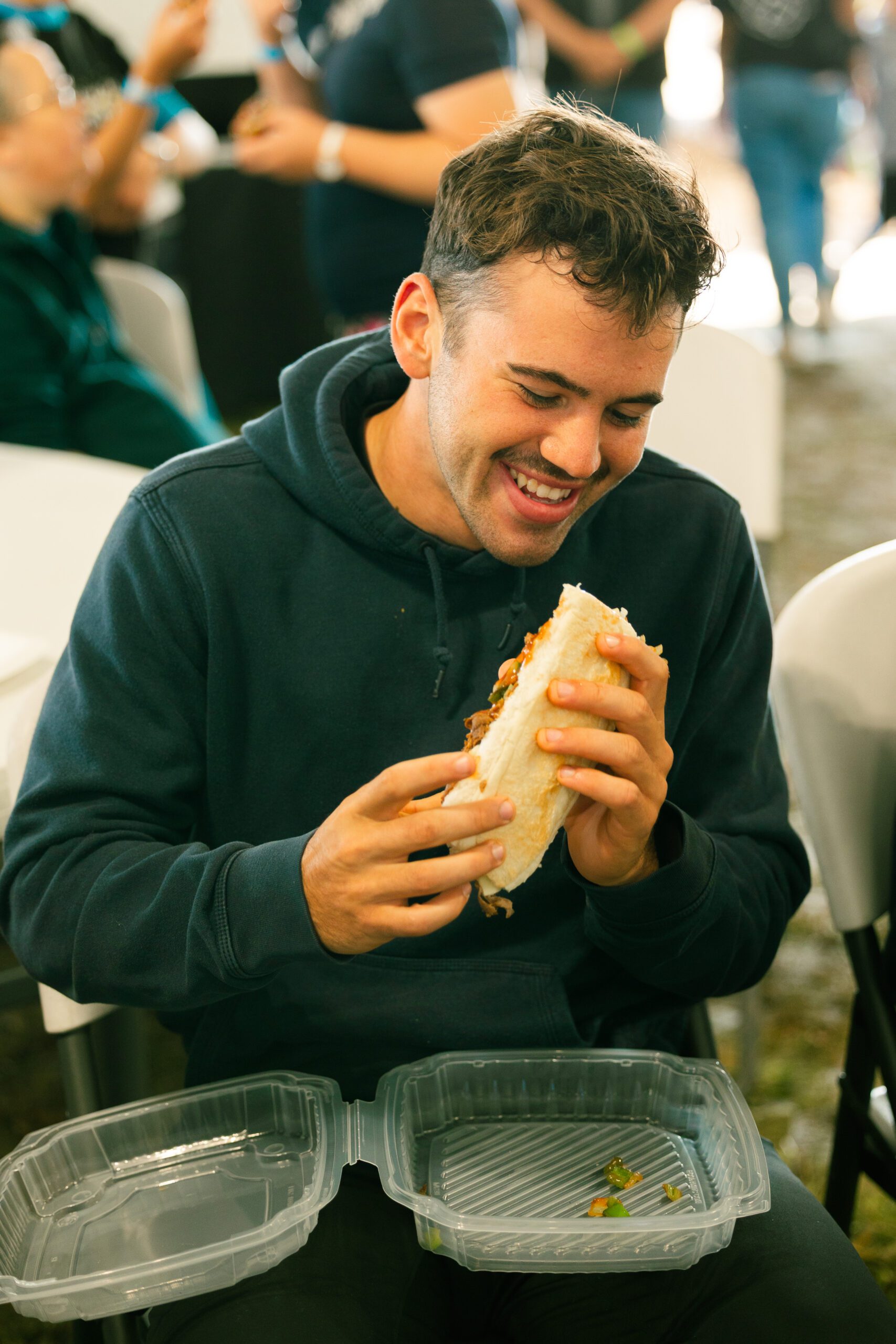A young man enjoying a large sandwich while seated at a table, with empty takeout containers in front of him, surrounded by a lively event atmosphere.