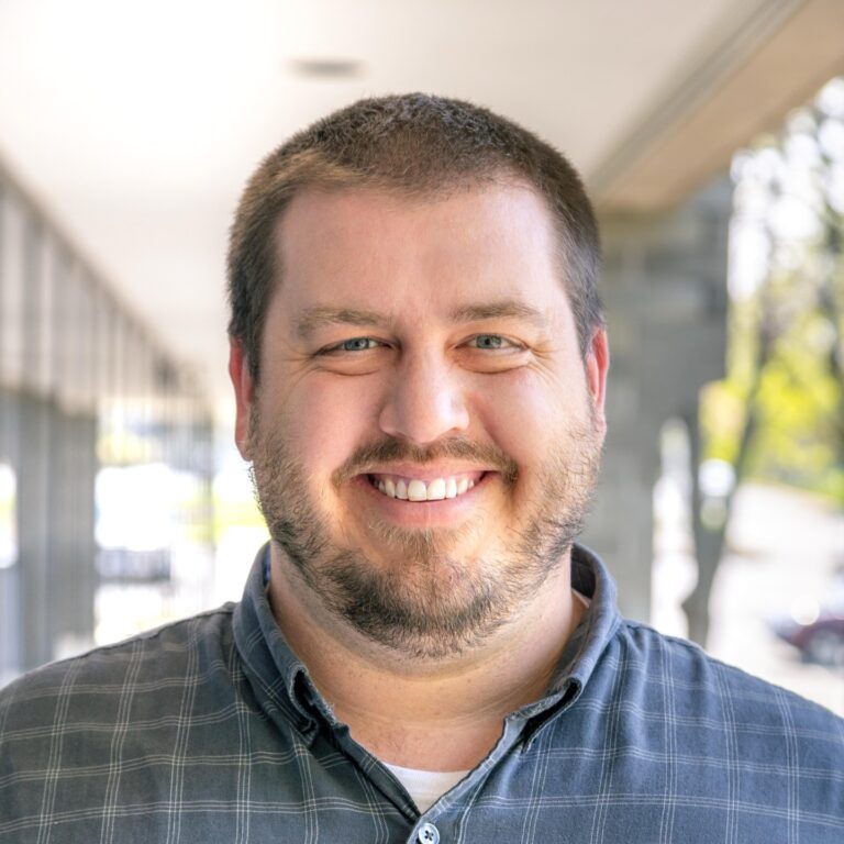 Smiling man with a beard wearing a plaid shirt, standing outdoors in a well-lit environment with blurred background elements.