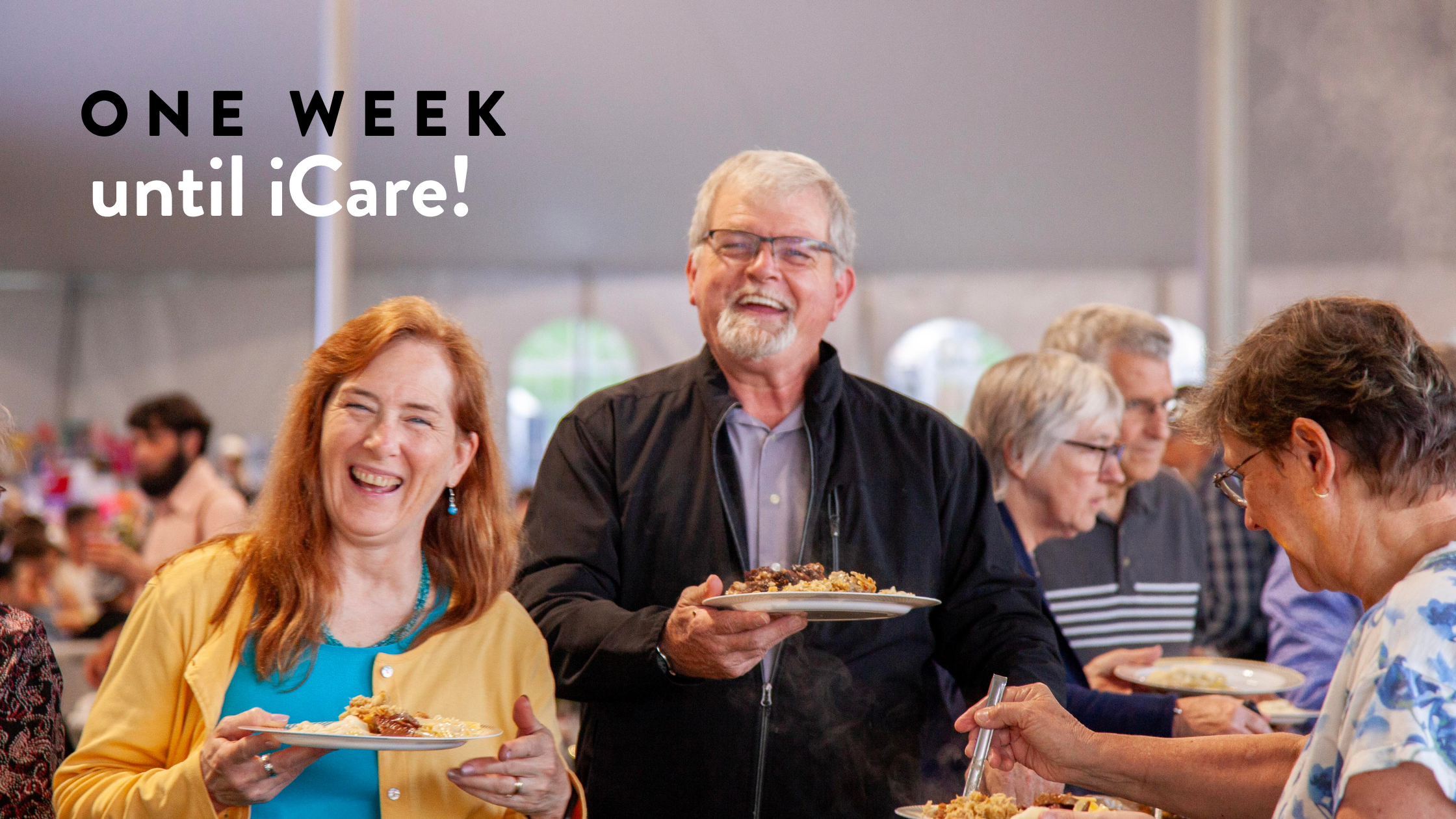 Group of smiling people enjoying a meal at a community event, with a banner stating "One Week until iCare!" in the background. The atmosphere is lively and inviting, showcasing a sense of togetherness and celebration.